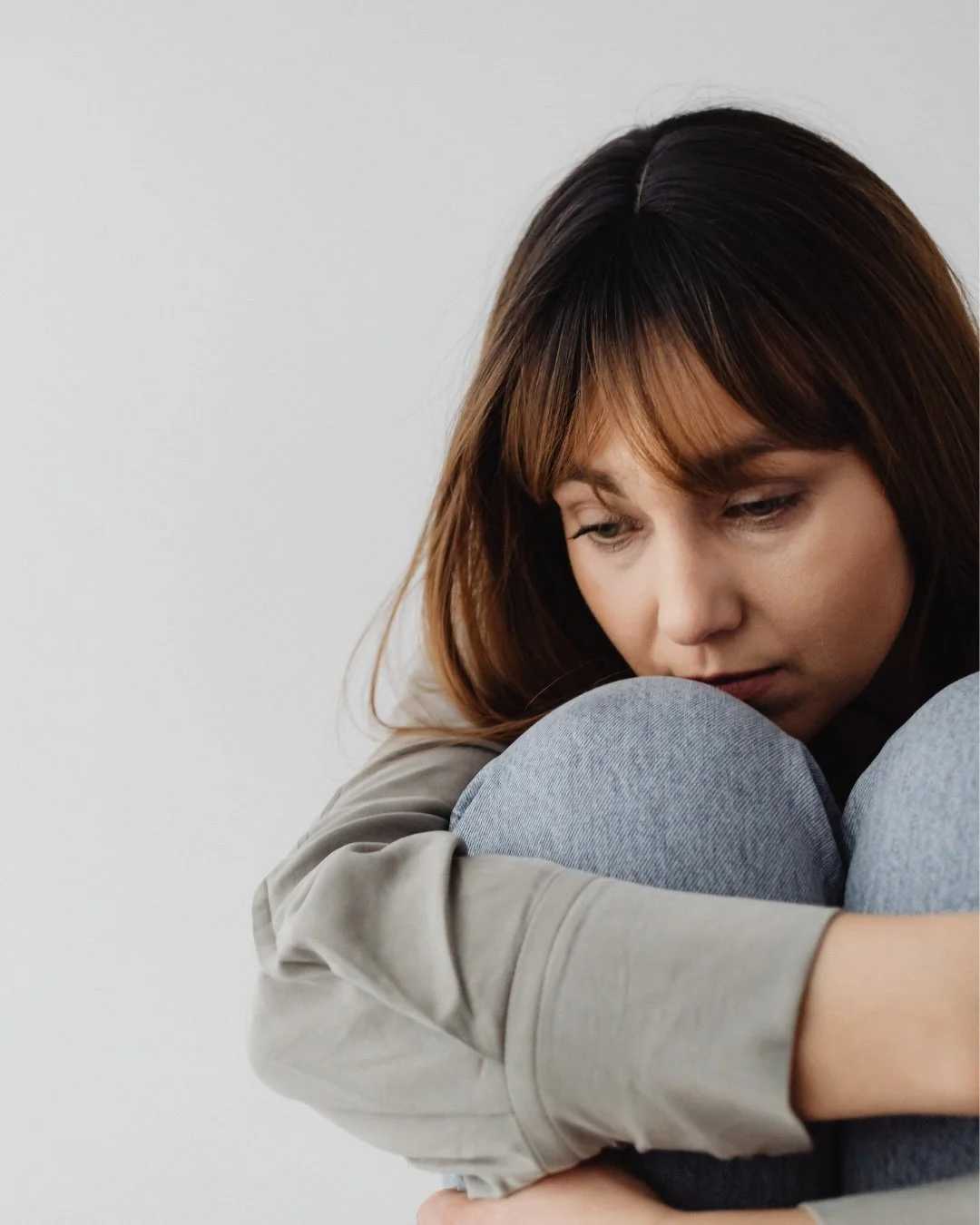 A woman with brown hair sitting on the floor hugging her knees, looking down with a sad expression.