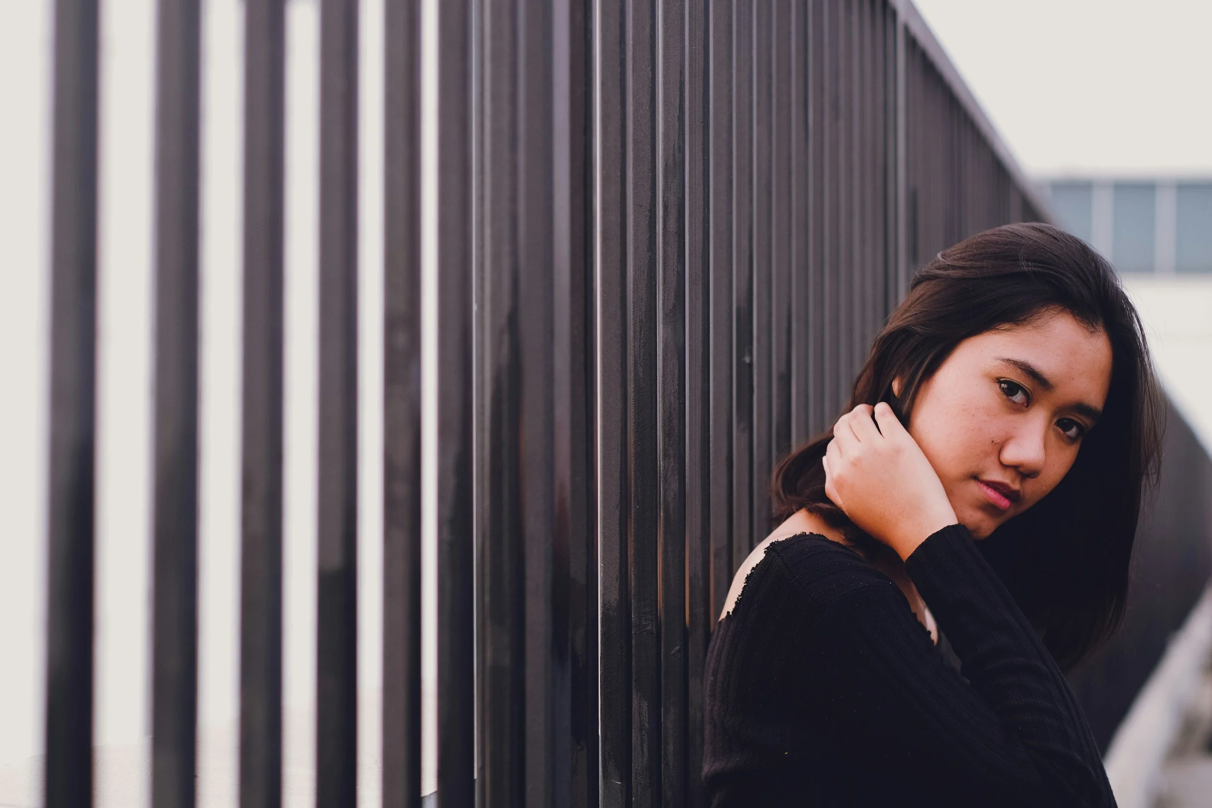 A young woman with dark hair and a black top leaning against a black metal fence, looking at the camera.