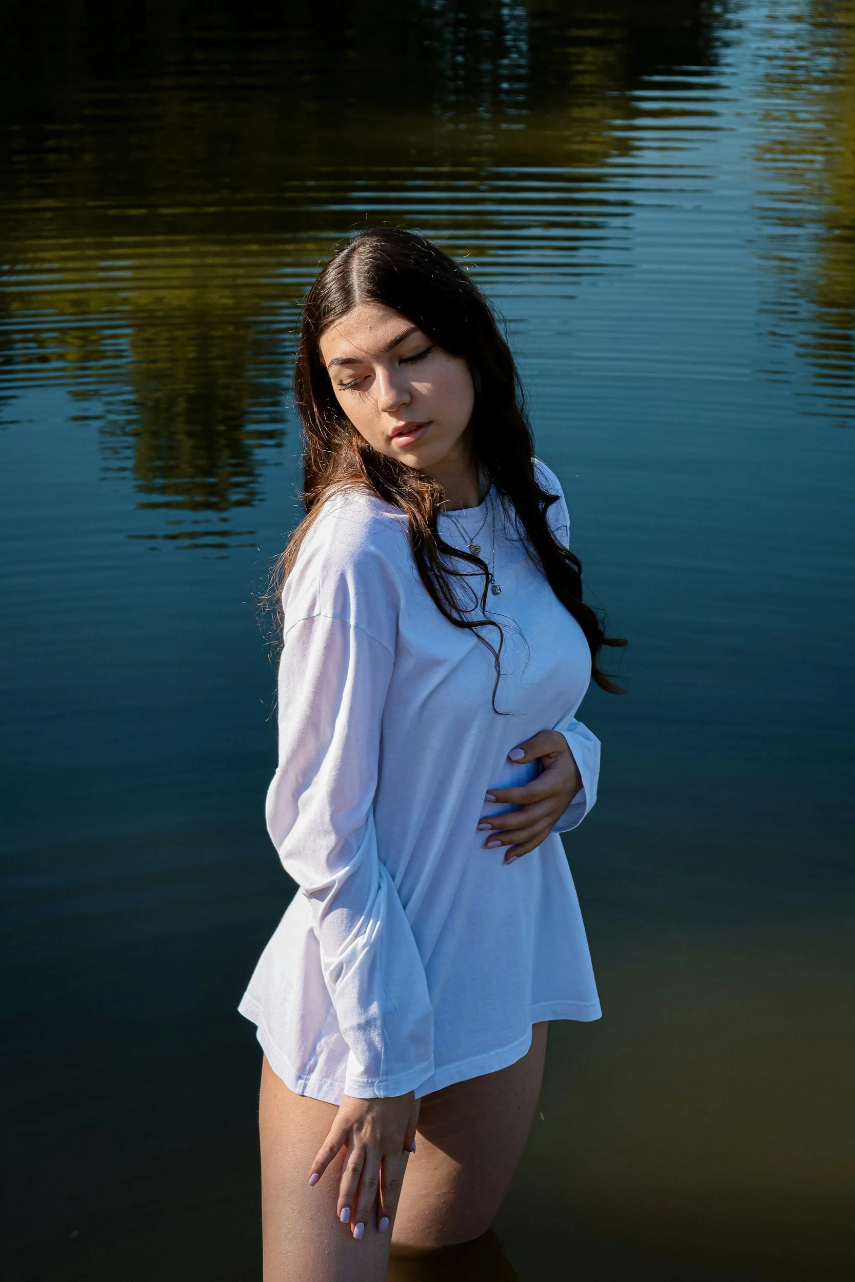 A young woman with long dark hair standing in a body of water, wearing a white long-sleeve shirt, with her eyes closed and one hand on her stomach, during daytime.