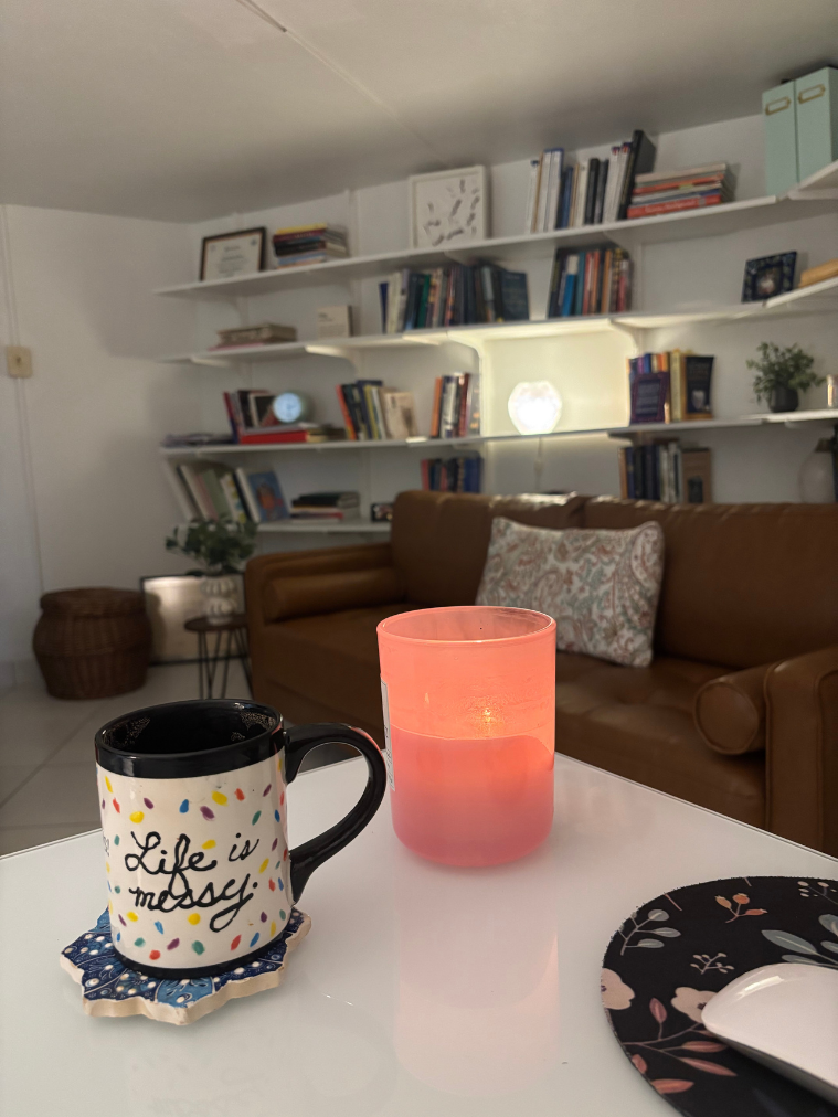 A cozy living room with a brown leather sofa, shelves filled with books, and a white table in the foreground. On the table are a decorated mug with the words 'Life is messy' and colorful dots, a pink candle holder, and a small patterned plate with a spoon.