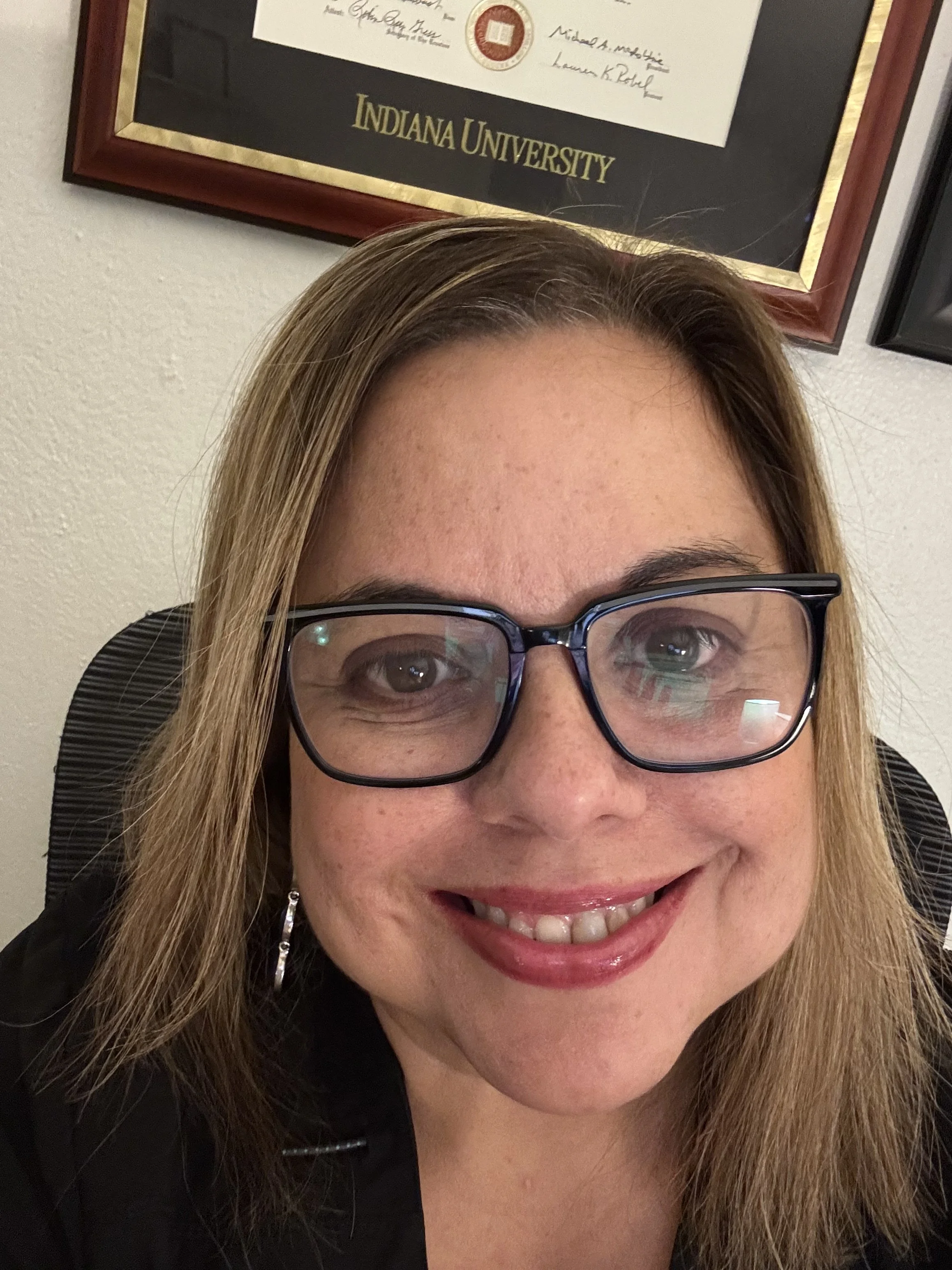 A woman with glasses and shoulder-length blonde hair smiling at the camera. Behind her, a framed diploma from Indiana University is visible on the wall.