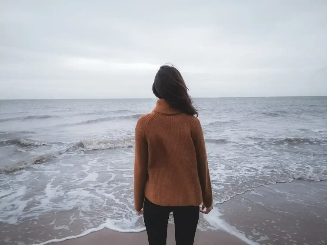 A person in a brown sweater standing on a beach facing the ocean with overcast skies.