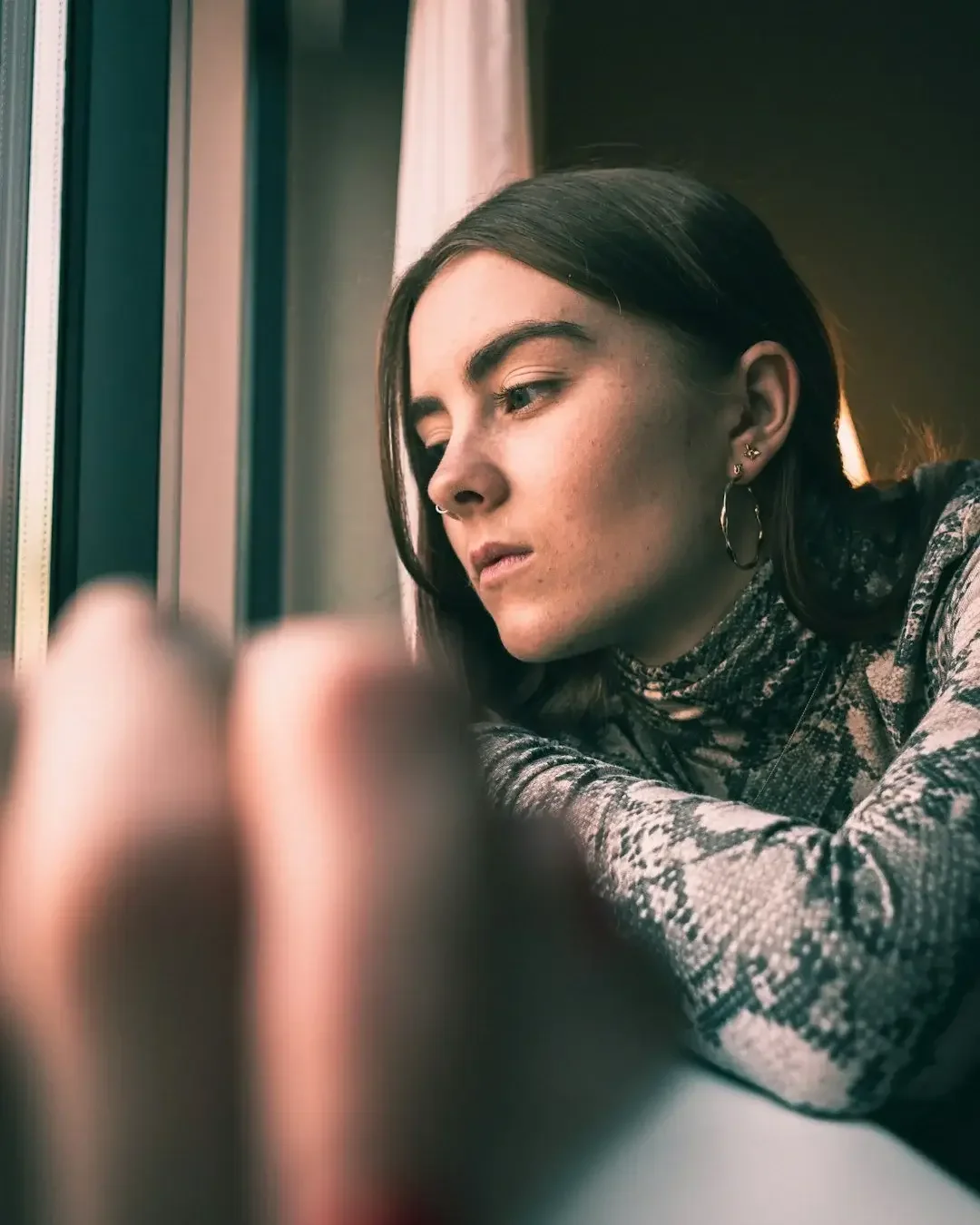 A young woman with dark hair, wearing earrings and a patterned top, looks out a window with a contemplative expression.