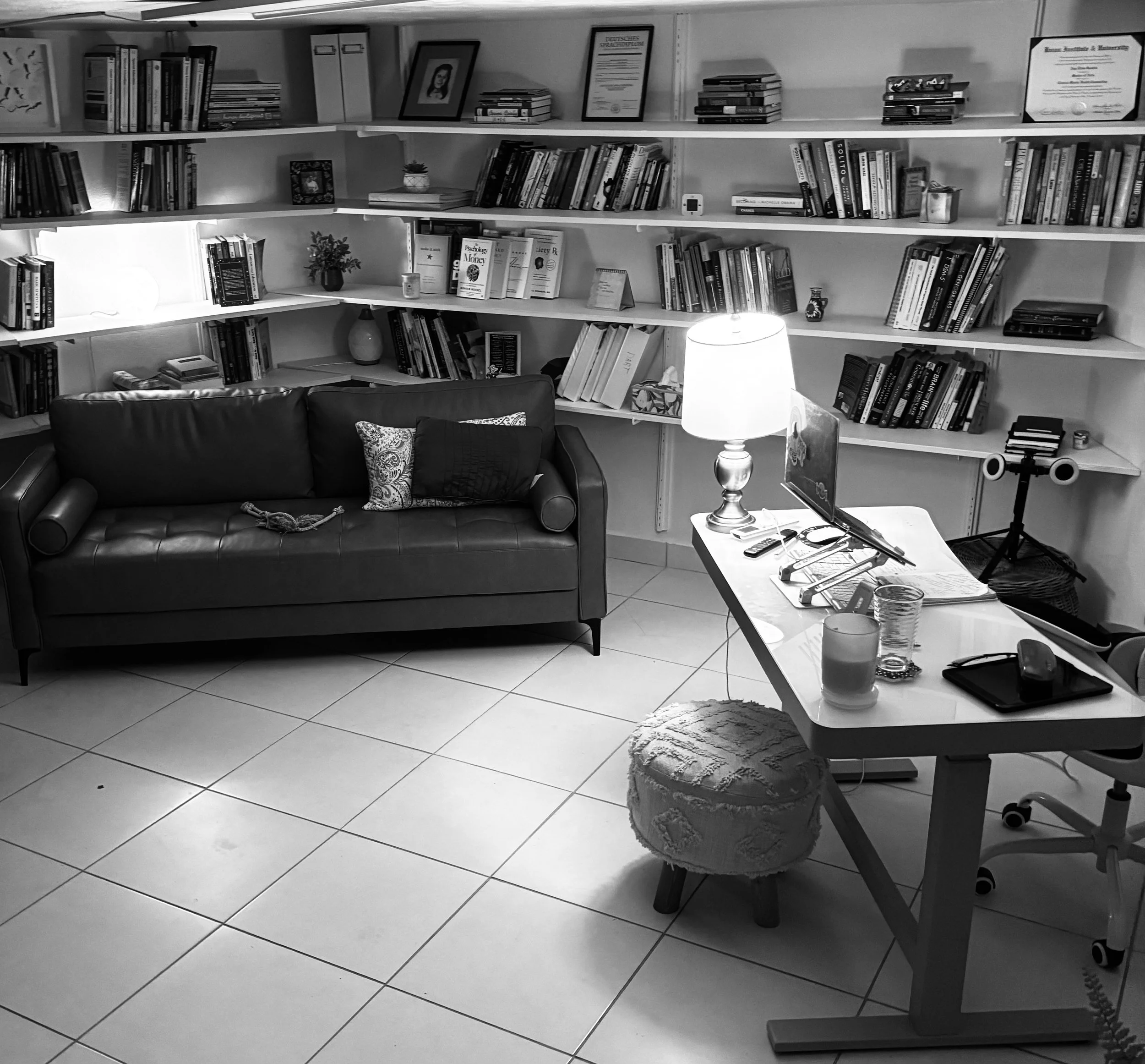 A cozy living room with a leather sofa, throw pillows, and a bookshelf filled with books and framed pictures. A desk with a lamp, laptop, and other items is next to the sofa, and a small, cushioned stool is in front of the desk.