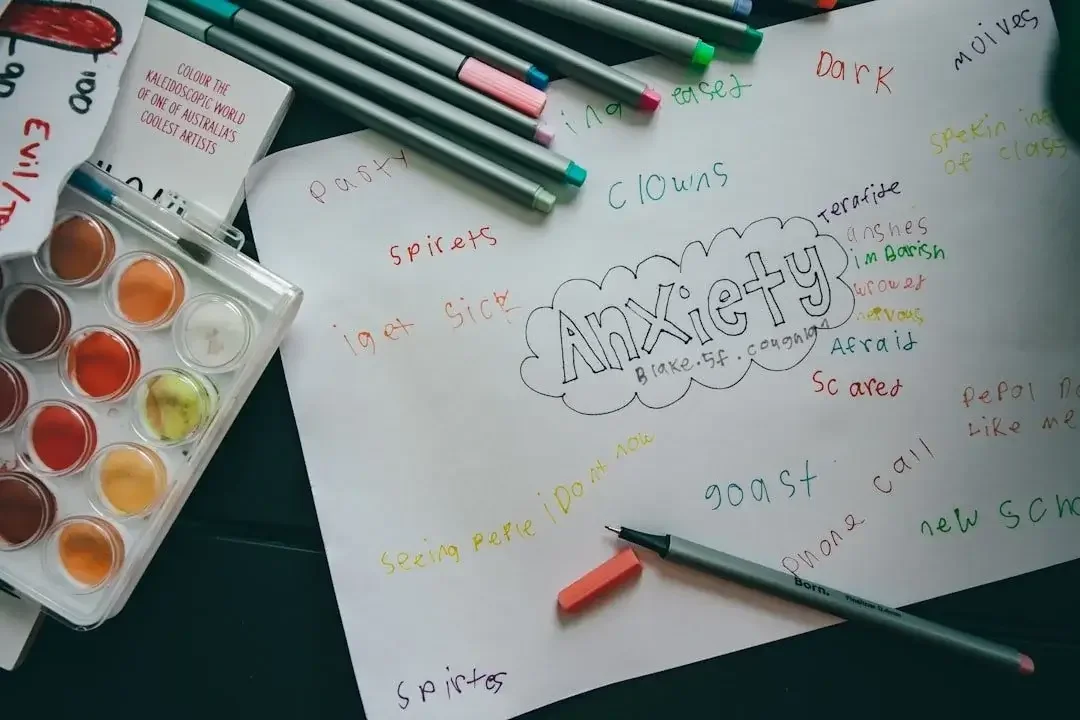 A handwritten list of words related to anxiety on a white paper, surrounded by colored markers, watercolor paints, and an eraser.
