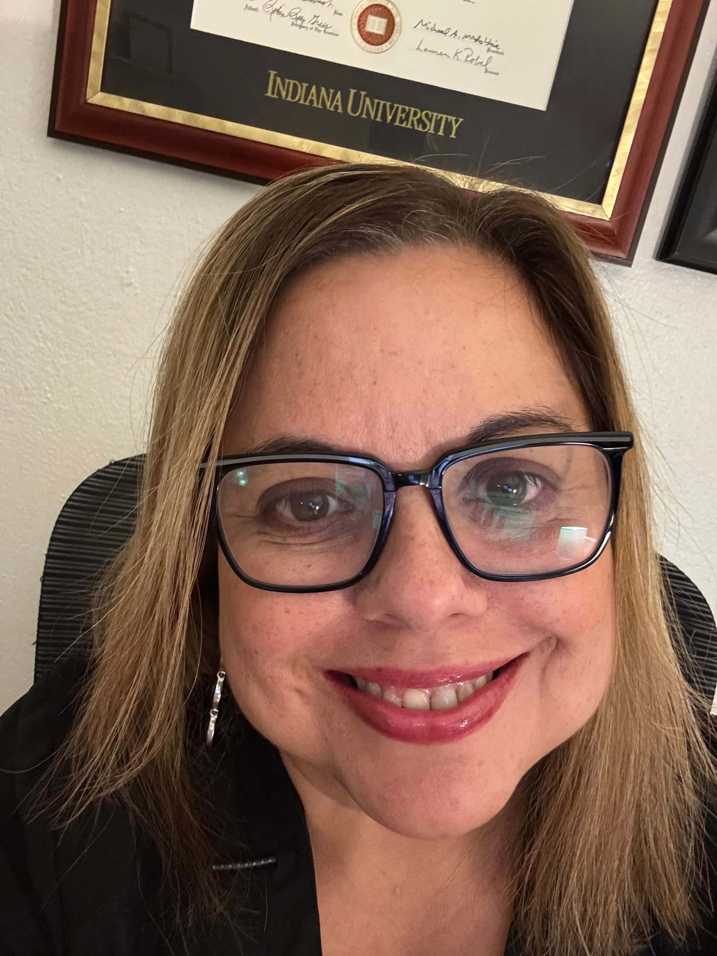Close-up of a smiling woman with long blonde hair, wearing glasses and a black top, with a framed Indiana University diploma on the wall behind her.