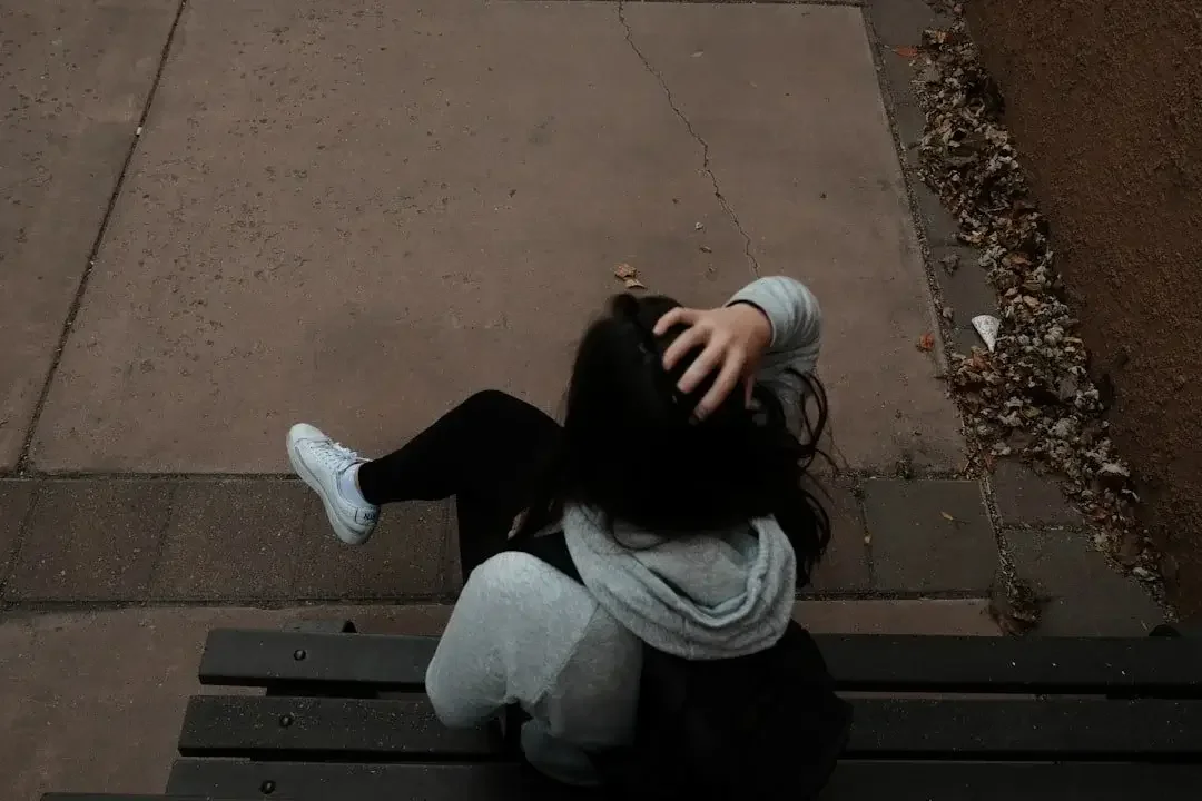 A person sitting on a park bench, with their hand on their head, wearing a gray hoodie and white sneakers, viewed from above.