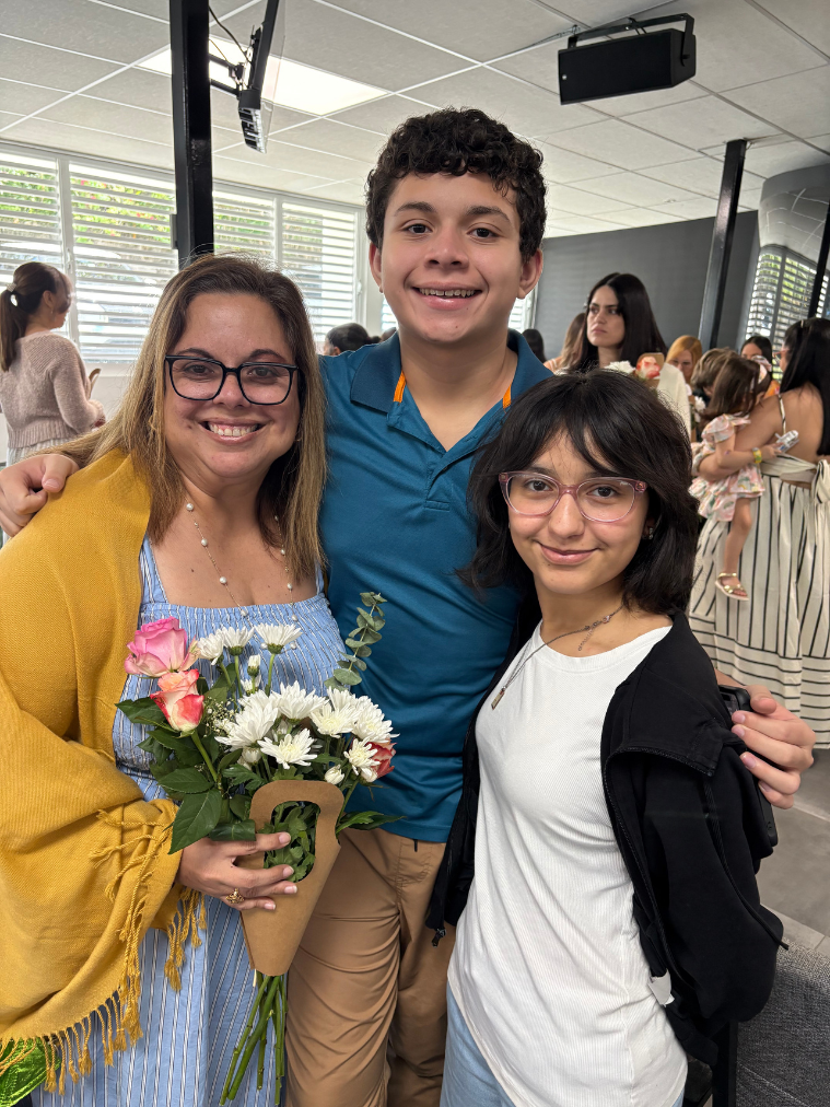 Three people smiling indoors, a woman holding a bouquet of flowers, a young man and a young woman, at a gathering or celebration event.