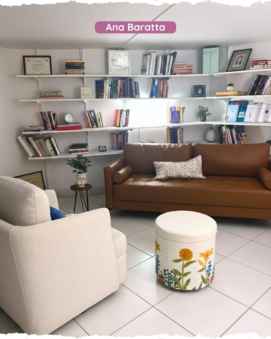A cozy living room with white tile floors, a beige armchair with a blue cushion, a brown leather sofa with decorative pillows, a small round ottoman with a floral pattern, and a wall of white shelves filled with books, framed pictures, plants, and decorative items.