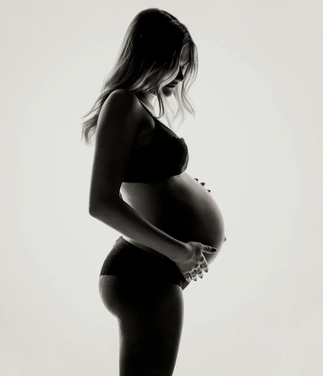 Silhouette of a pregnant woman in black lingerie with her hands on her belly, against a plain light background.
