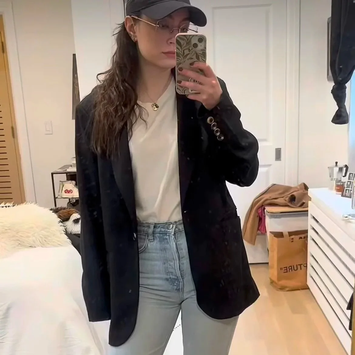 A woman taking a mirror selfie in a bedroom, wearing a black blazer, white t-shirt, light jeans, baseball cap, and glasses, with a smartphone with a floral case.