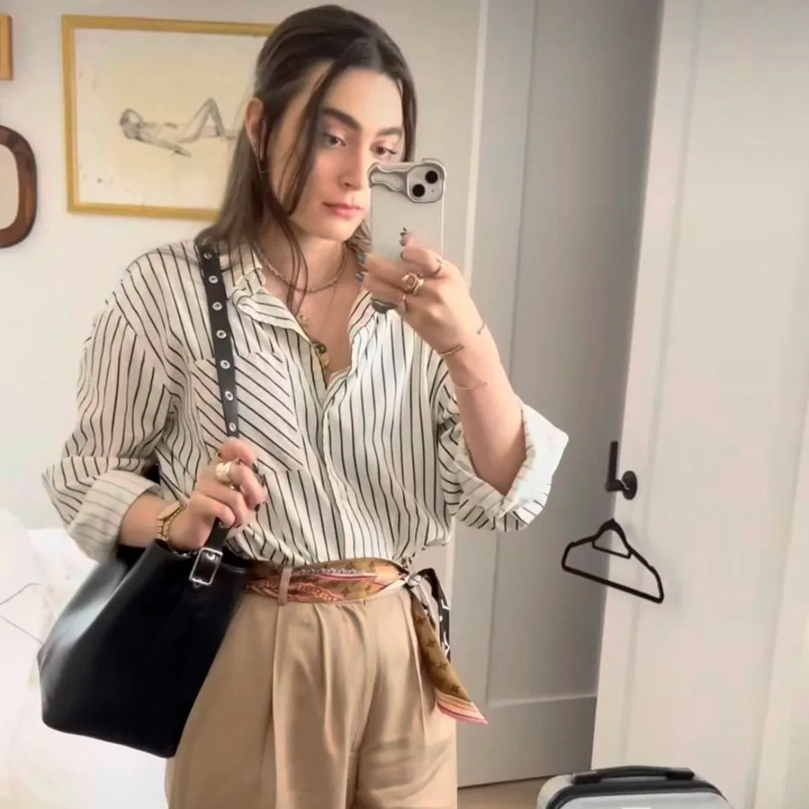 A young woman taking a mirror selfie in a room, wearing a striped blouse, beige pants, and carrying a black handbag.