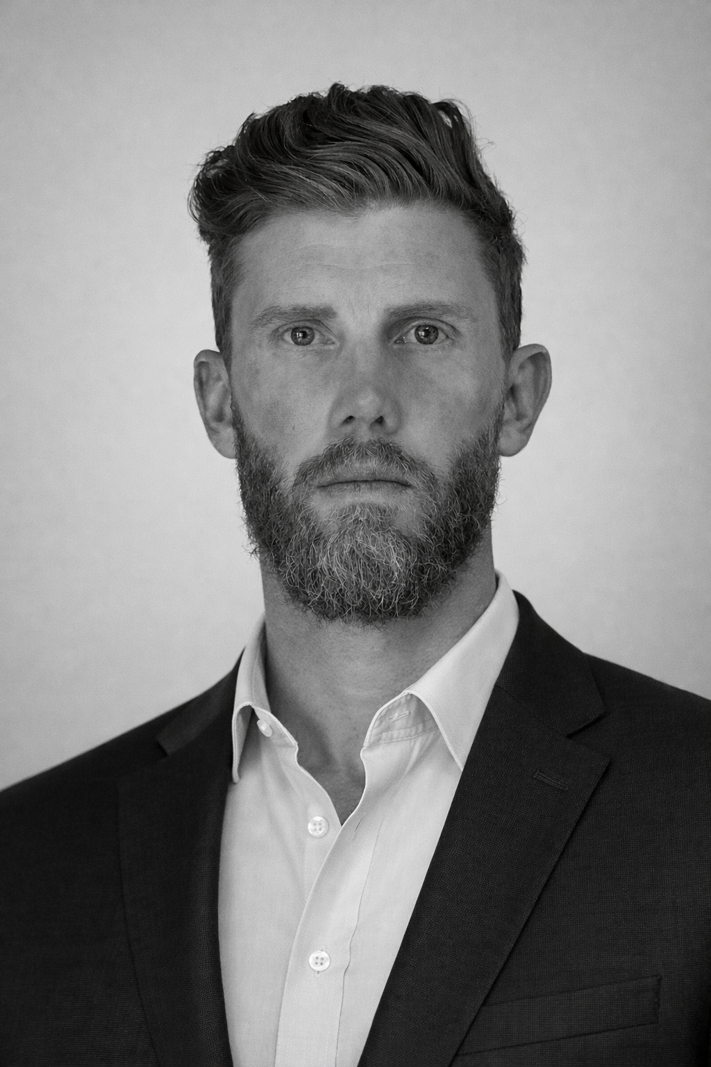 Black and white portrait of a man with a beard and serious expression, wearing a suit jacket and white shirt.