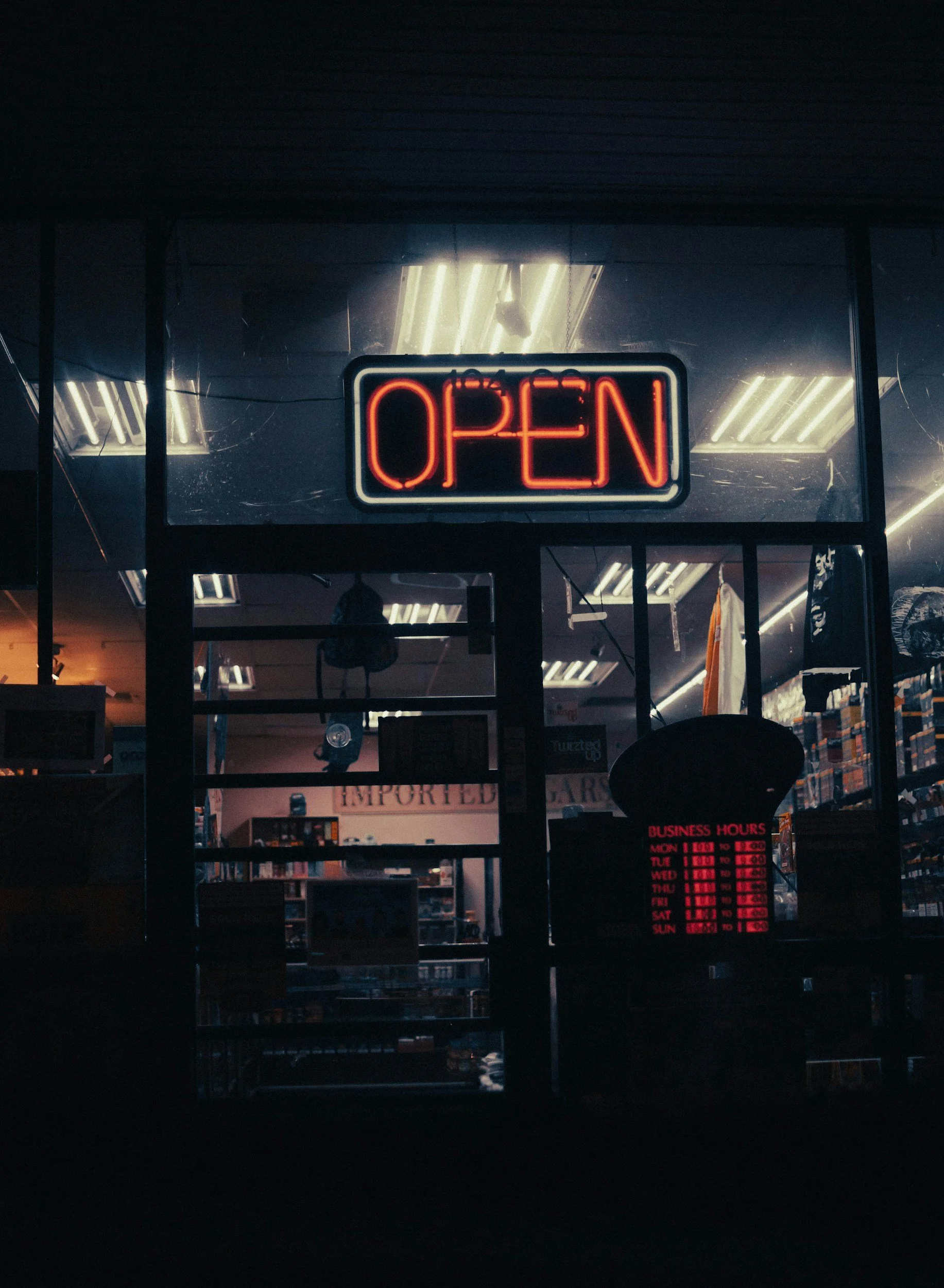 Neon 'OPEN' sign outside a store at night with business hours displayed.