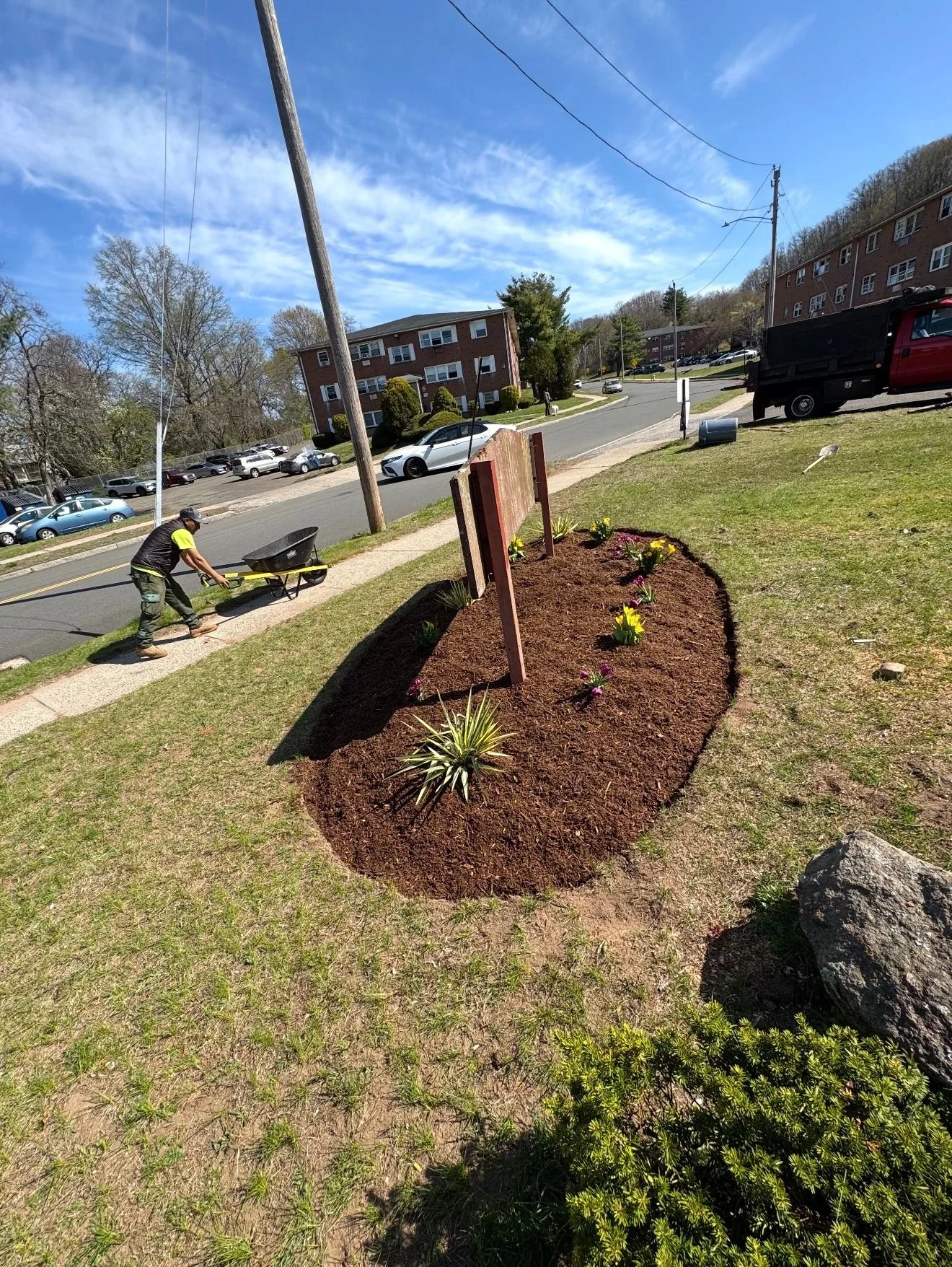 Person pushing a wheelbarrow along sidewalk near a freshly planted flower bed in a grassy area on a sunny day.