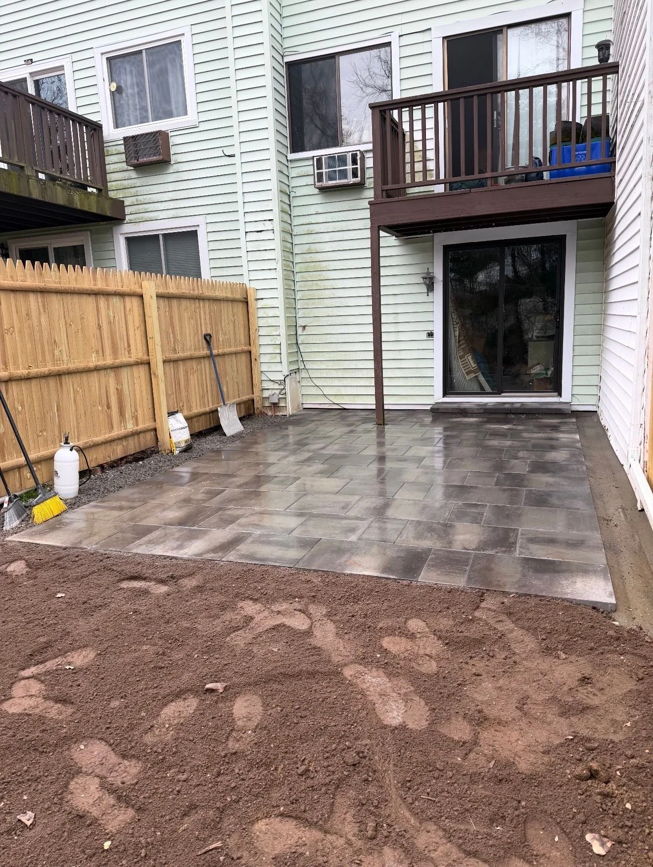 Backyard patio with fresh stone tiles, a wooden fence on the left, and a sliding glass door leading into the house. There are tools and a spray bottle leaning against the fence, and a small second-floor balcony with patio furniture is visible.