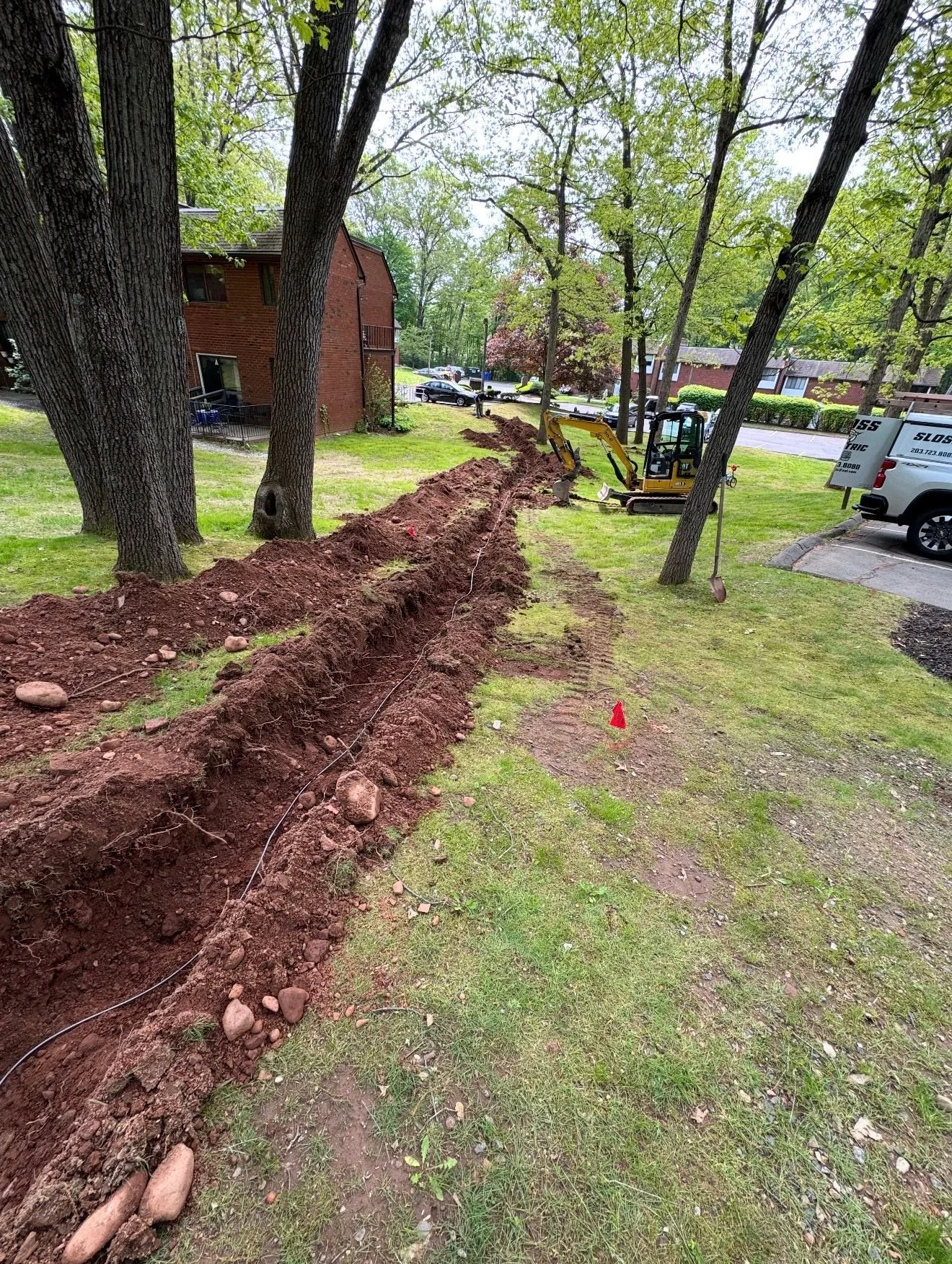 A trench is being dug in a grassy area next to trees and a residential building with construction equipment nearby.