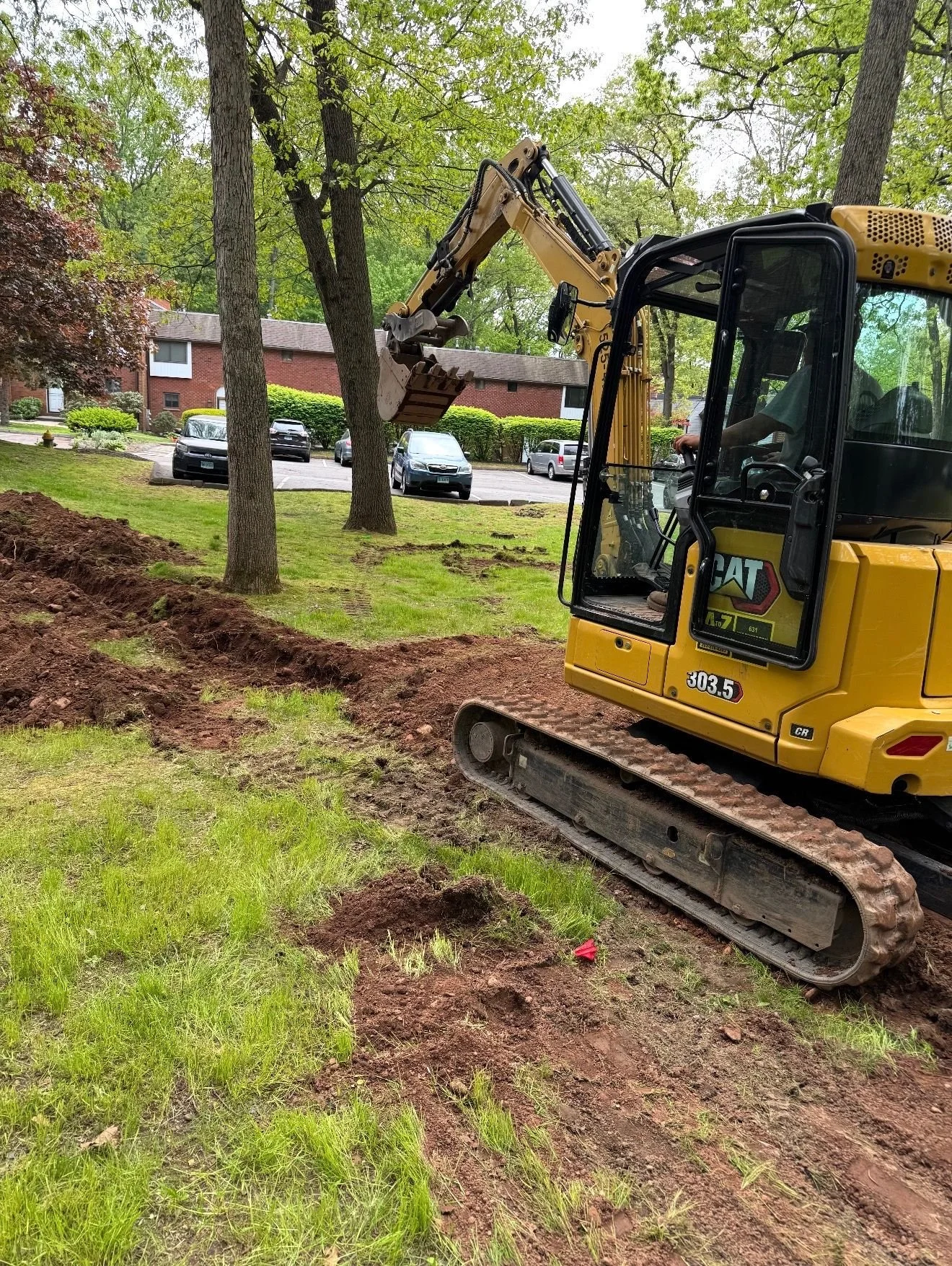 A yellow CAT excavator is digging a trench on a grassy area with trees and apartment buildings in the background.
