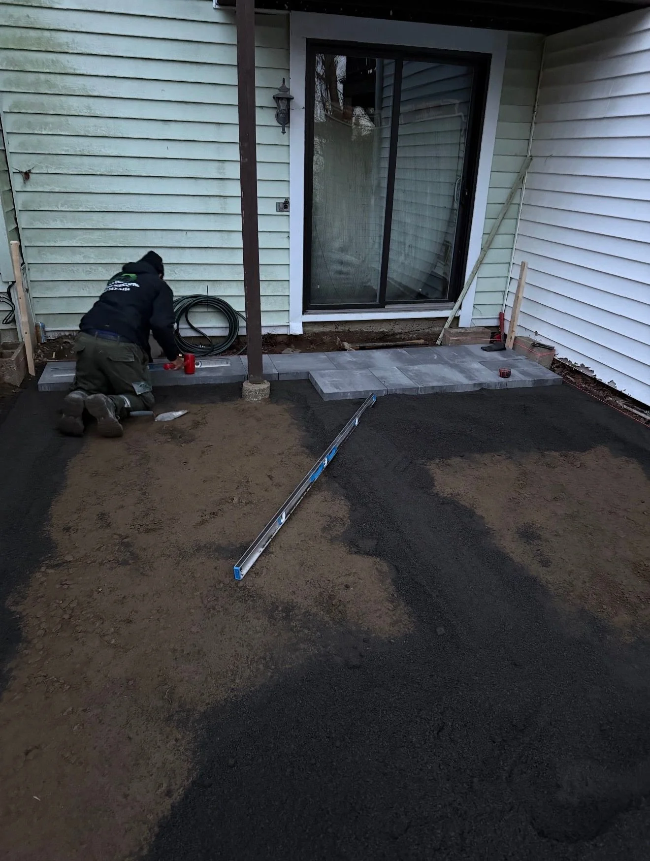 A worker installing outdoor paving stones next to a house, with a partially laid stone pathway, some tools, and a level on the ground.