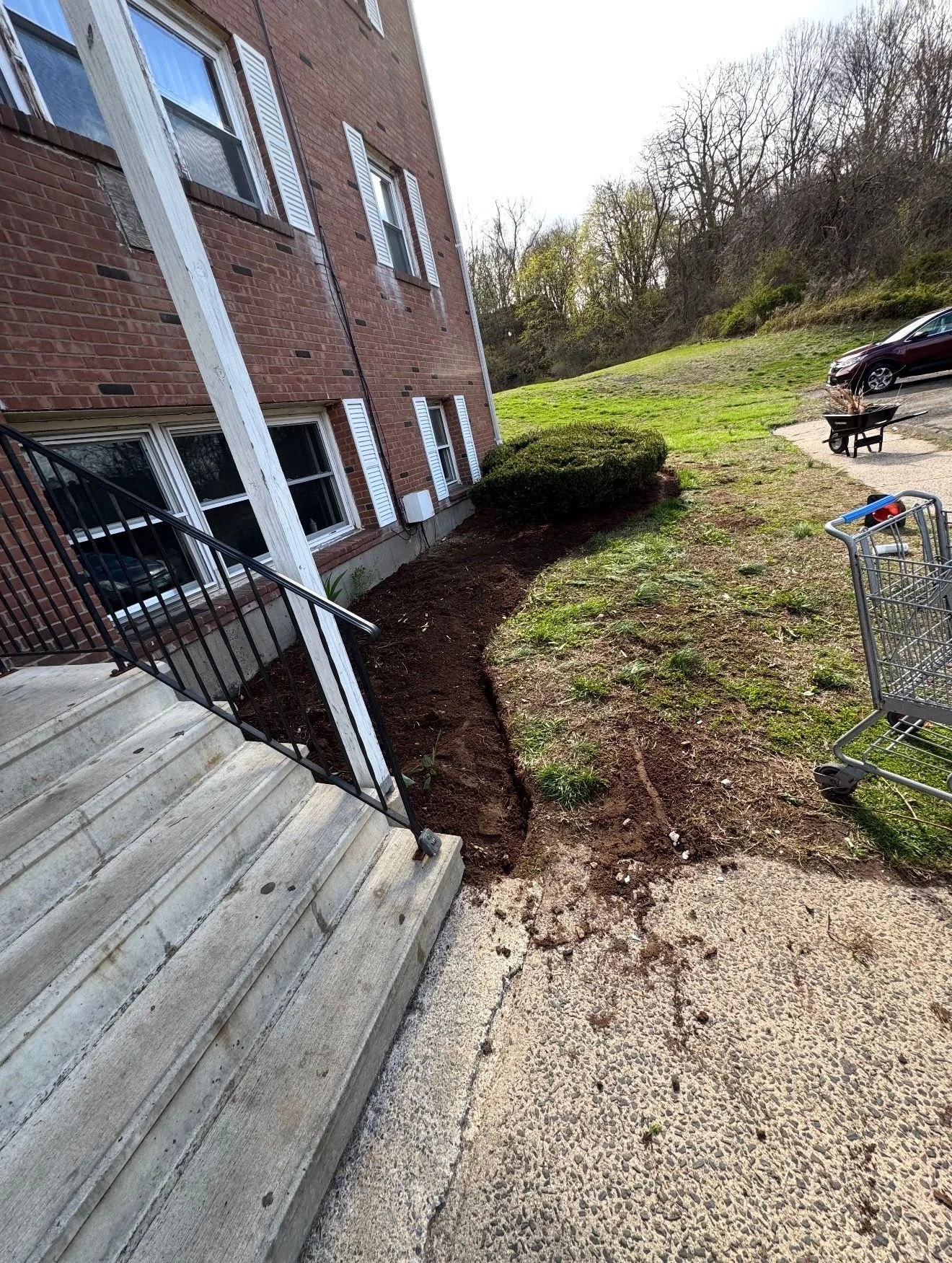 View of the side of a red brick apartment building with stairs, a black railing, and a partially dug garden bed along the sidewalk. There is a bush and a grassy area with a car, a wheelbarrow, and a shopping cart nearby.