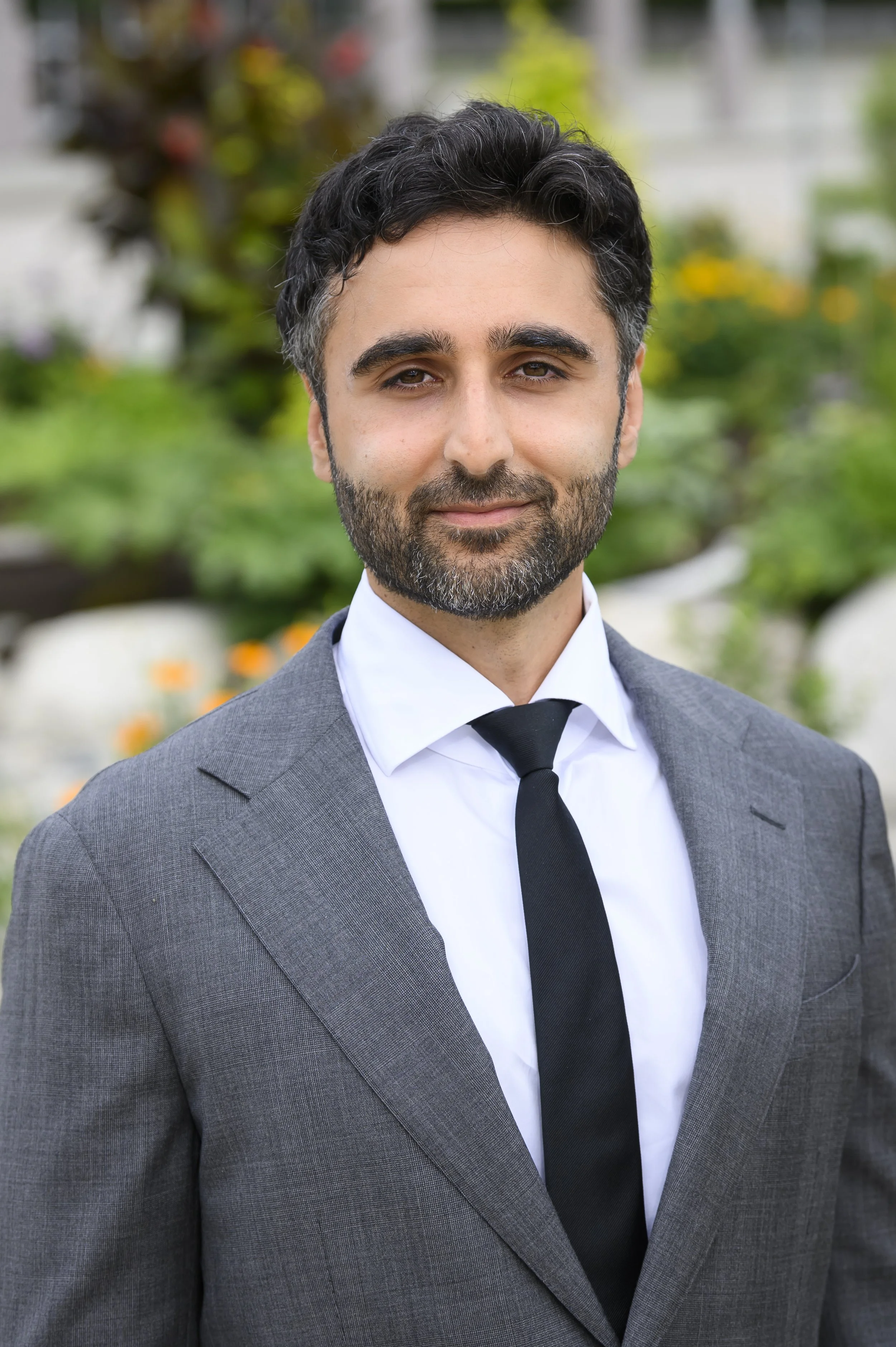 A man with dark, curly hair and a beard wearing a gray suit, white shirt, and black tie, standing outdoors with greenery and colorful flowers in the background.