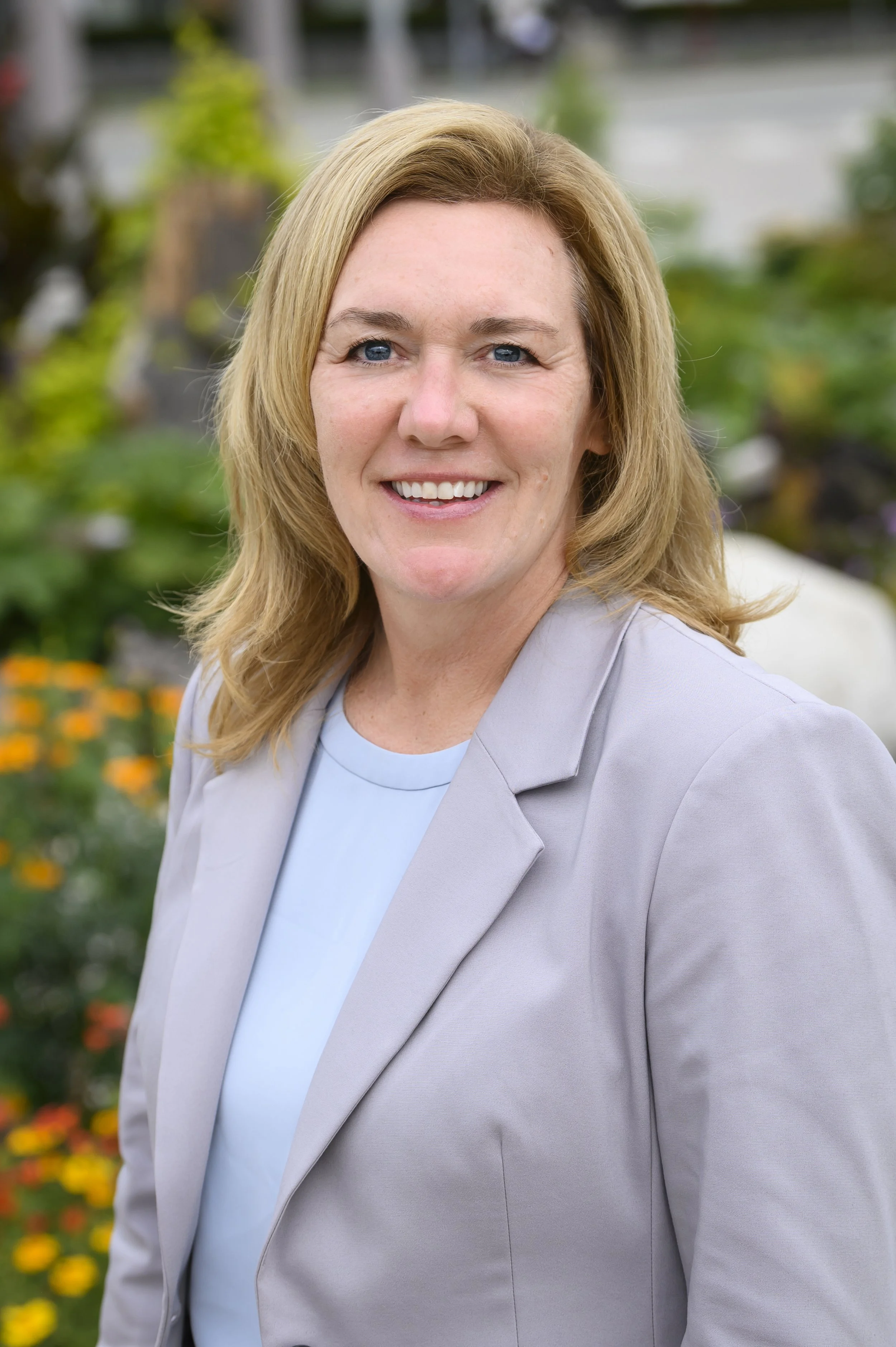 A smiling woman with blonde hair wearing a light-colored blazer and a light blue top, outdoors with blurred greenery and flowers in the background.