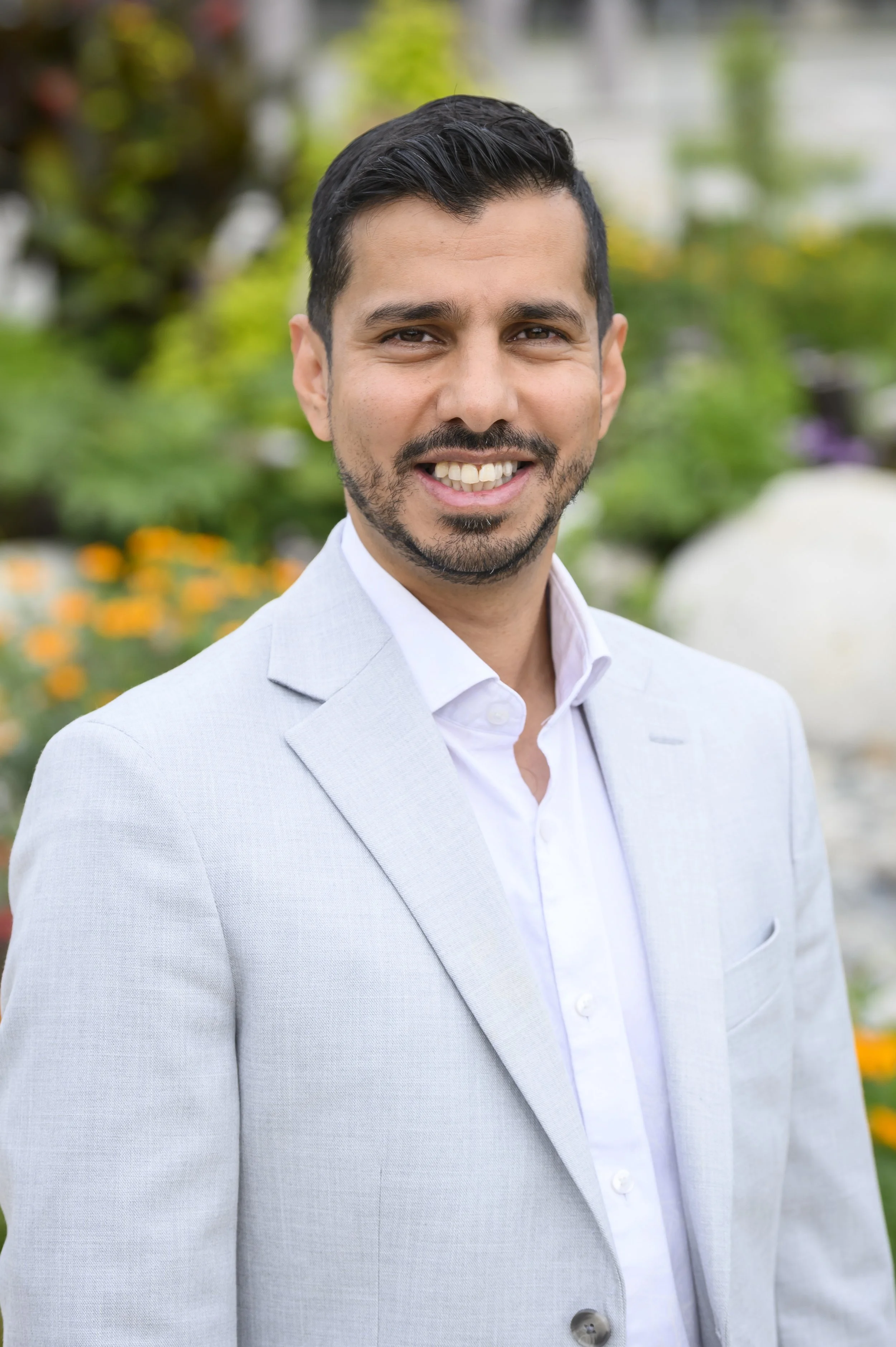 A man with dark hair and a beard wearing a light gray suit and white shirt smiles outdoors, with blurred greenery and flowers in the background.