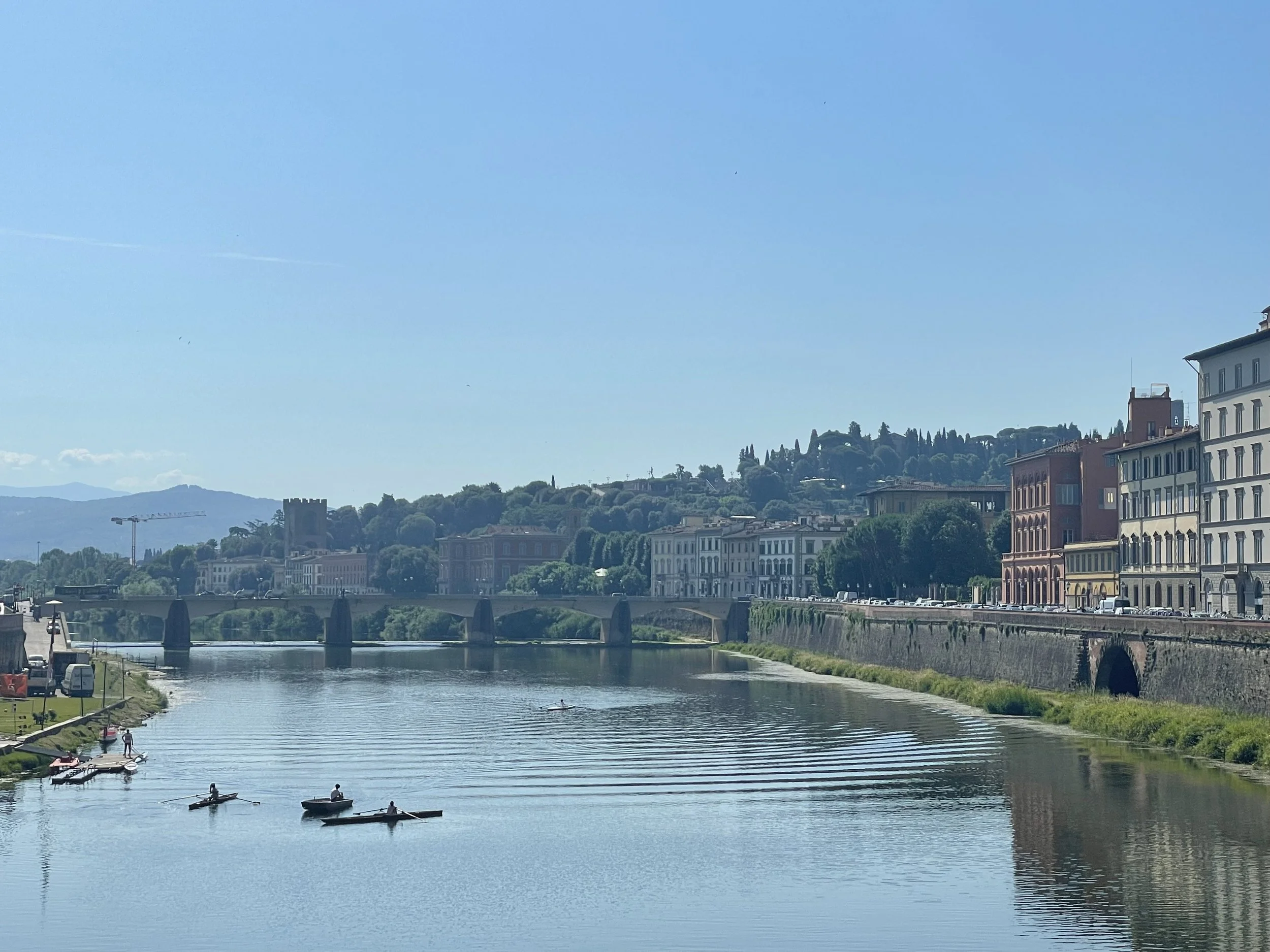 A scenic view of a river with small boats, a bridge, and historic buildings along the riverbank on a sunny day.