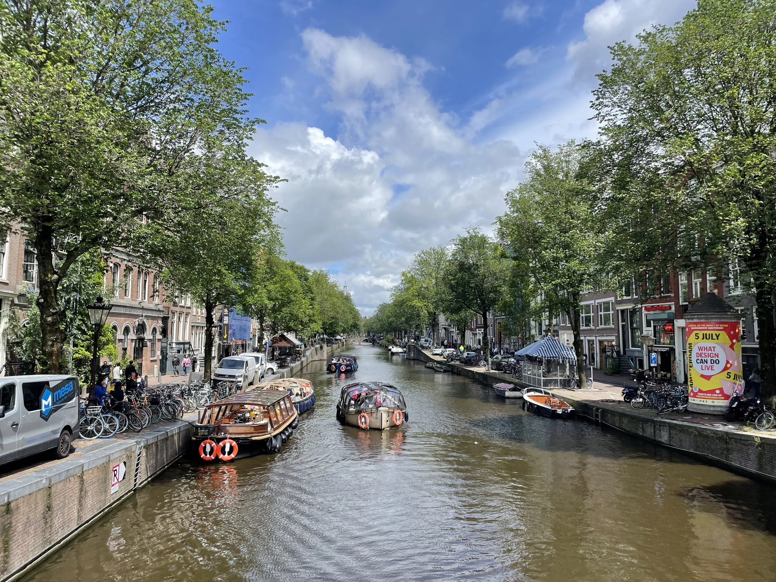 A canal scene in a European city, likely Amsterdam, with boats on the water, trees lining the edges, and buildings on both sides. The sky is partly cloudy.