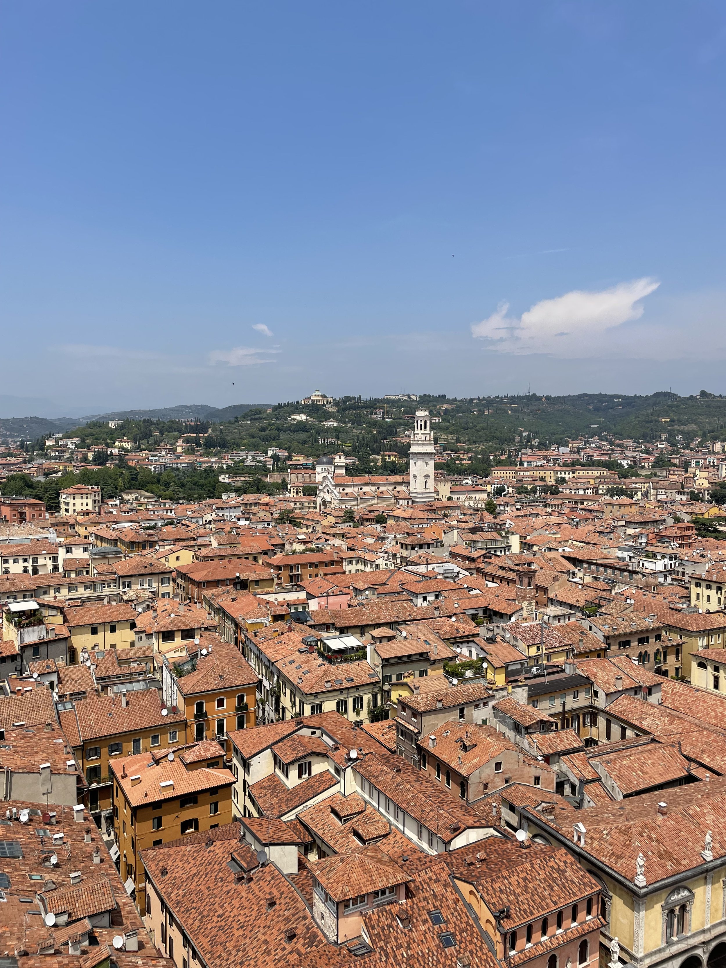A panoramic view of an Italian city with terracotta rooftops, a tall bell tower, and distant green hills under a partly cloudy sky.