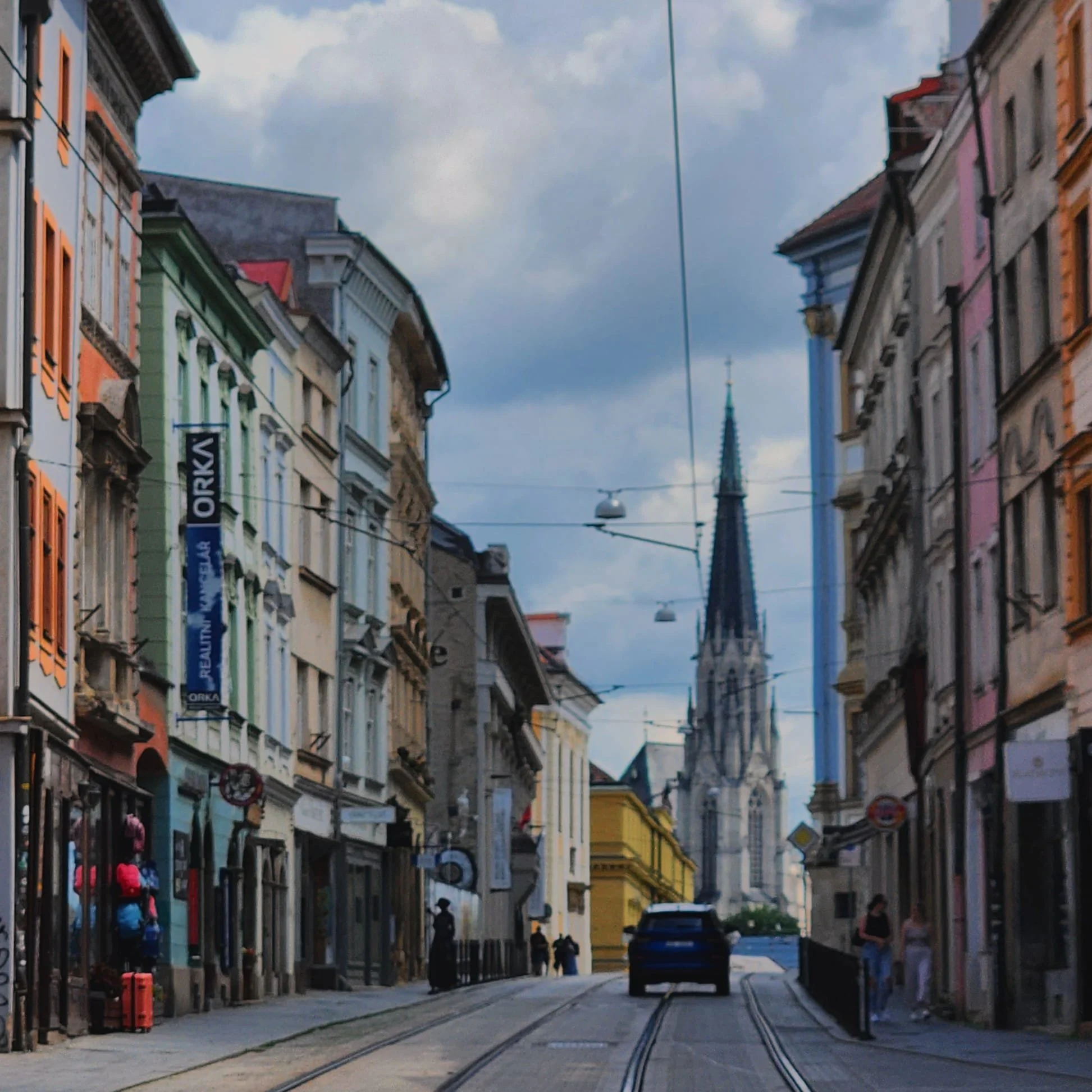 A city street with colorful buildings, tram tracks, and a prominent church steeple in the distance under a cloudy sky.