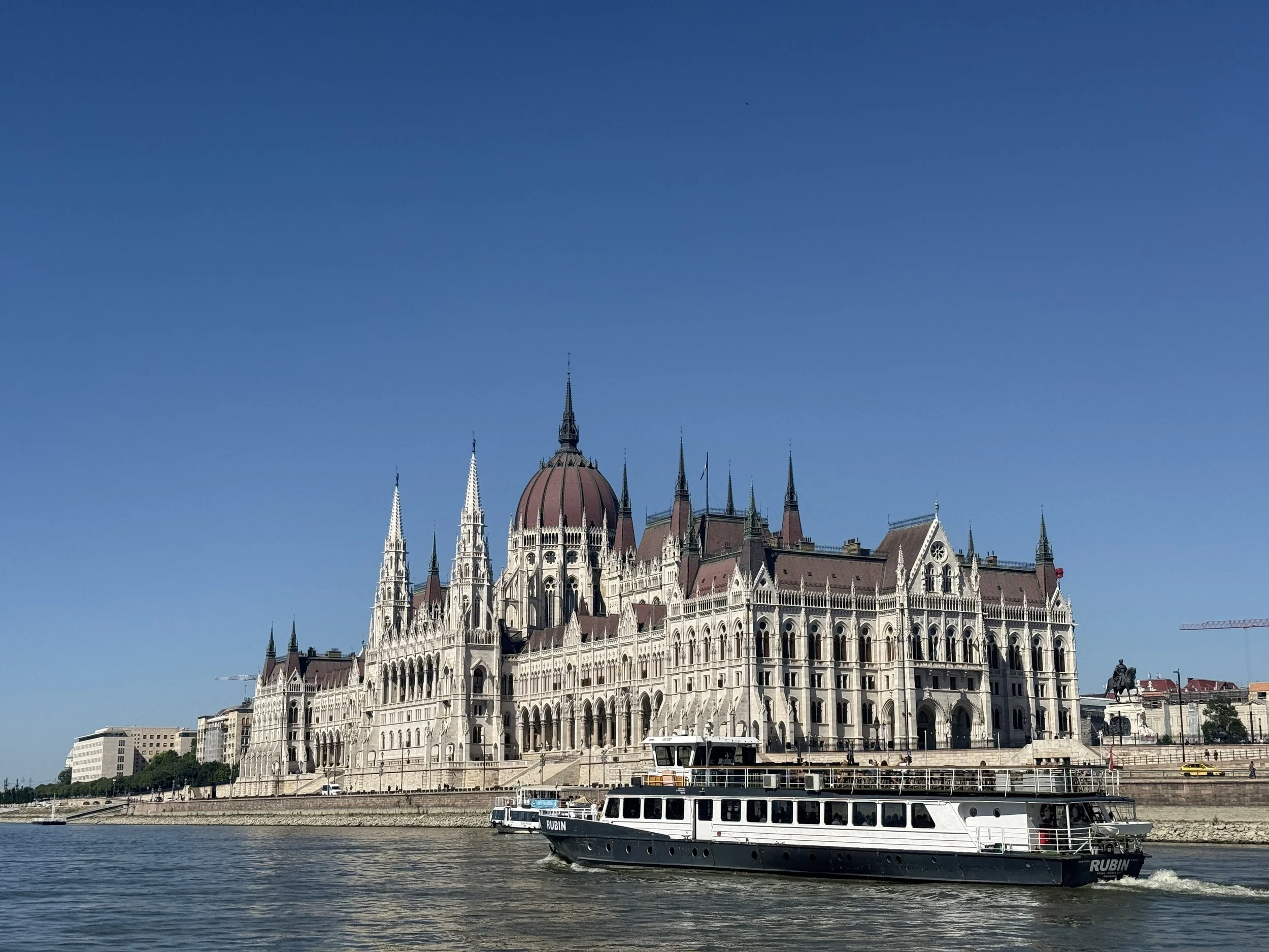 A historic castle-like building with gothic architecture along a river, with a boat named RUBIN sailing in the foreground under a clear blue sky.