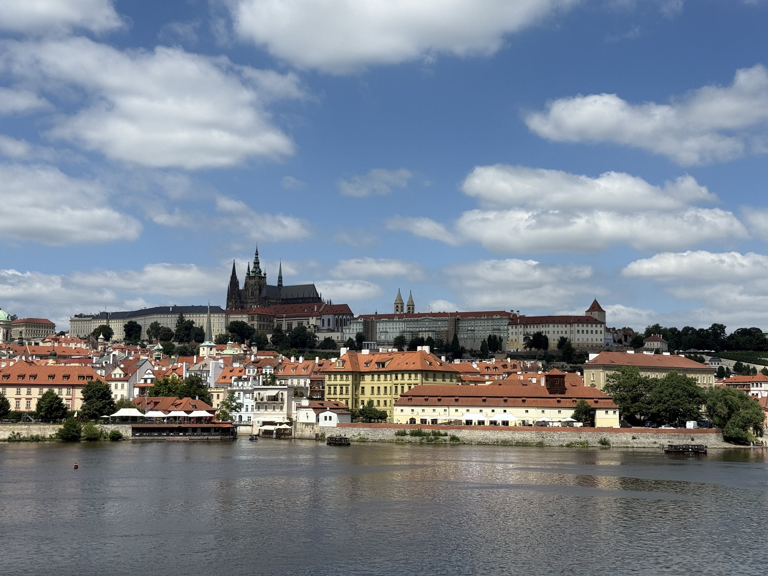 A panoramic view of a European cityscape with historic buildings along a river, featuring a castle or cathedral on a hill under a partly cloudy sky.