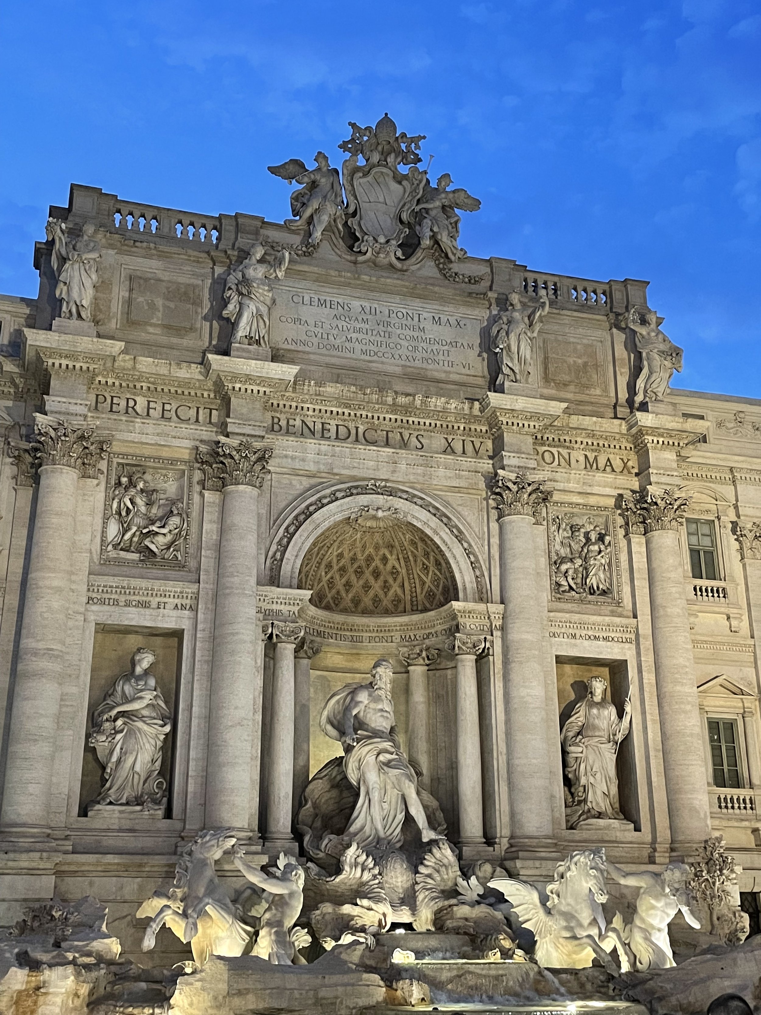 The Trevi Fountain in Rome, Italy, with ornate sculptures and a central figure, illuminated at dusk against a blue sky.