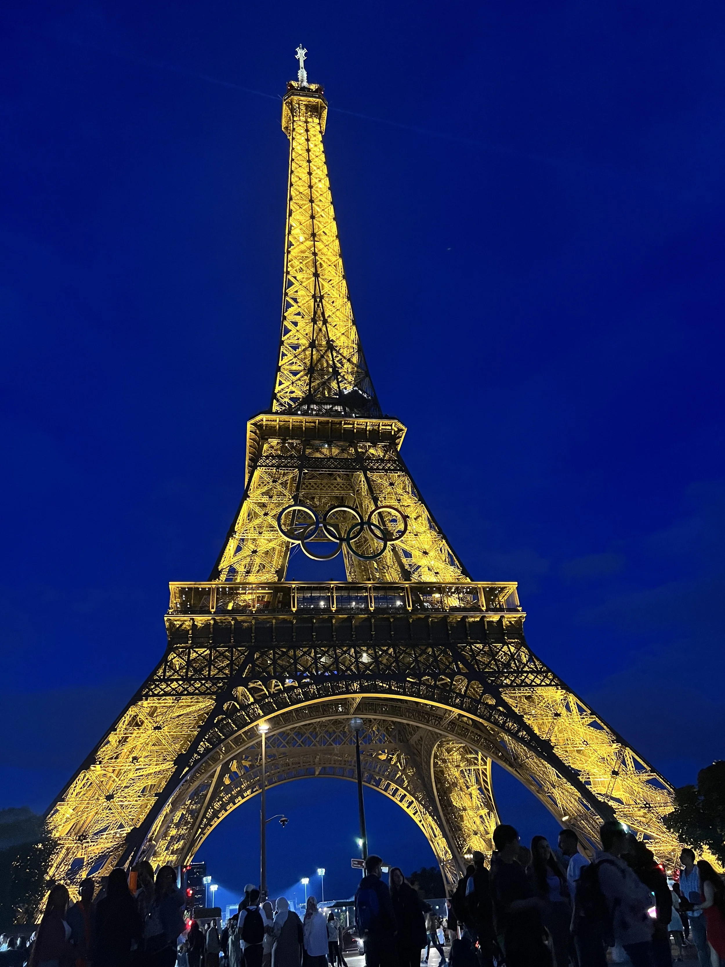 Eiffel Tower illuminated at night with the Olympic rings display and people gathering around.
