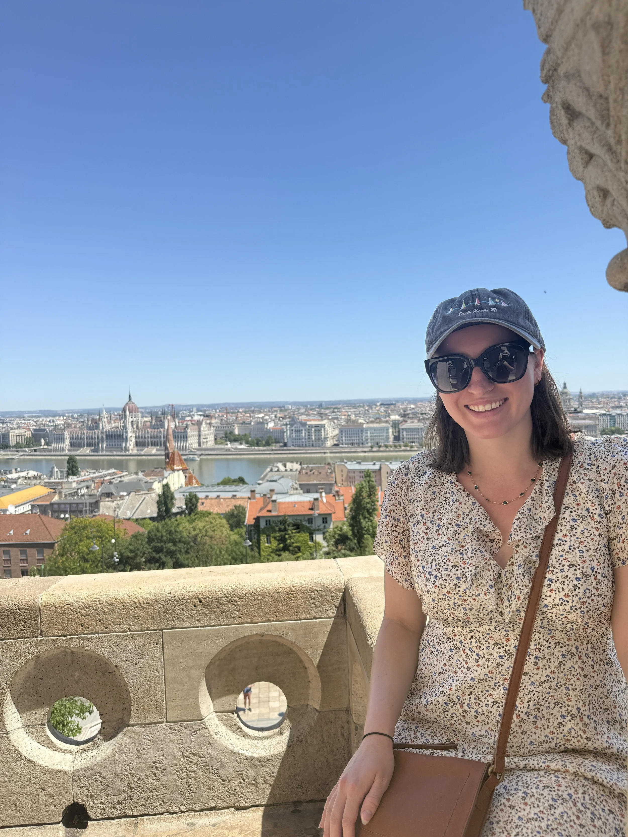 A woman wearing sunglasses, a cap, and a floral dress sitting on a historic stone balcony with a panoramic city view including the Parliament building and Danube River in Budapest, Hungary.