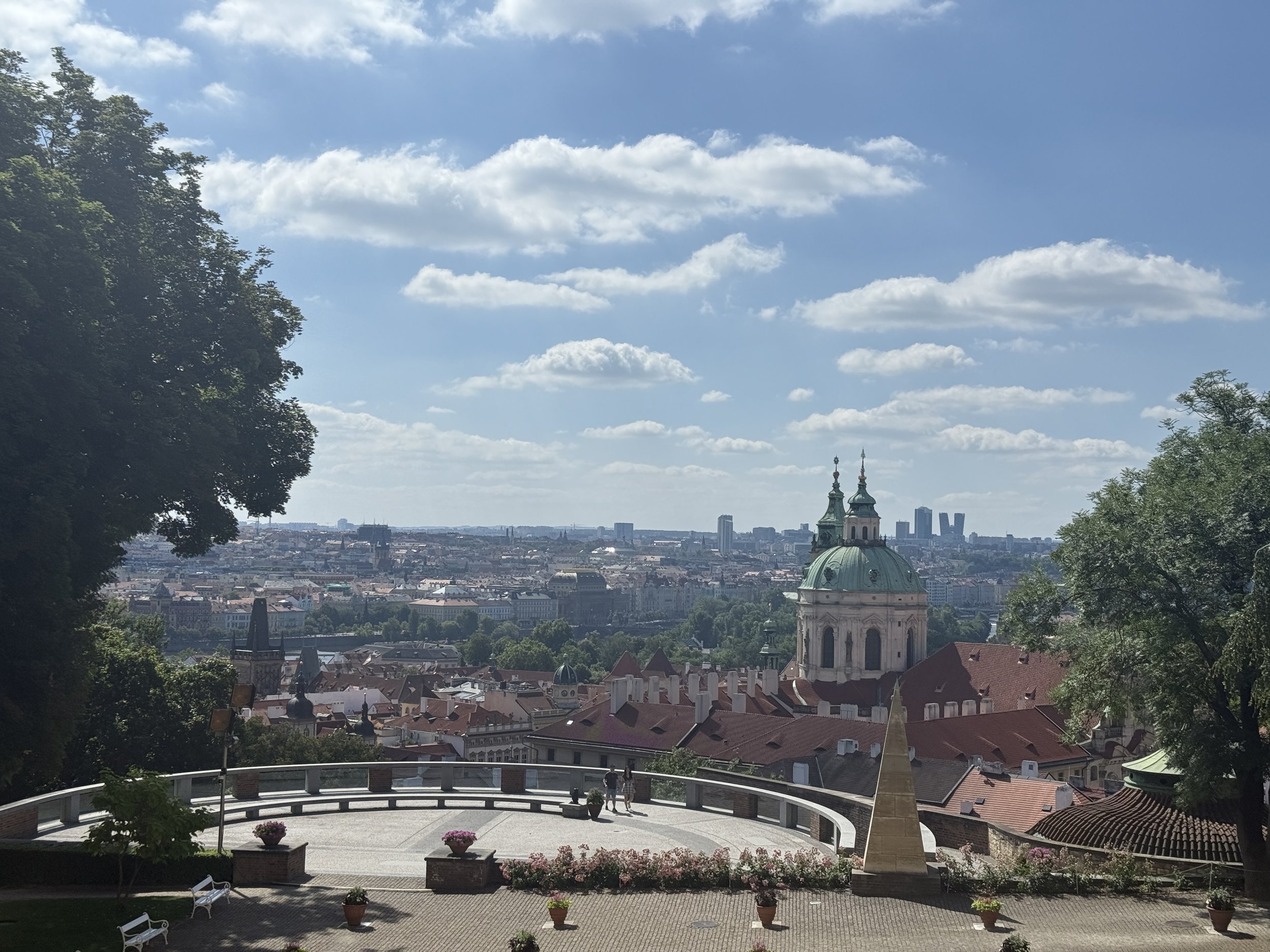 View of a cityscape with a church featuring a green dome and spires, seen from an elevated terrace with trees and flower pots, under a partly cloudy sky.