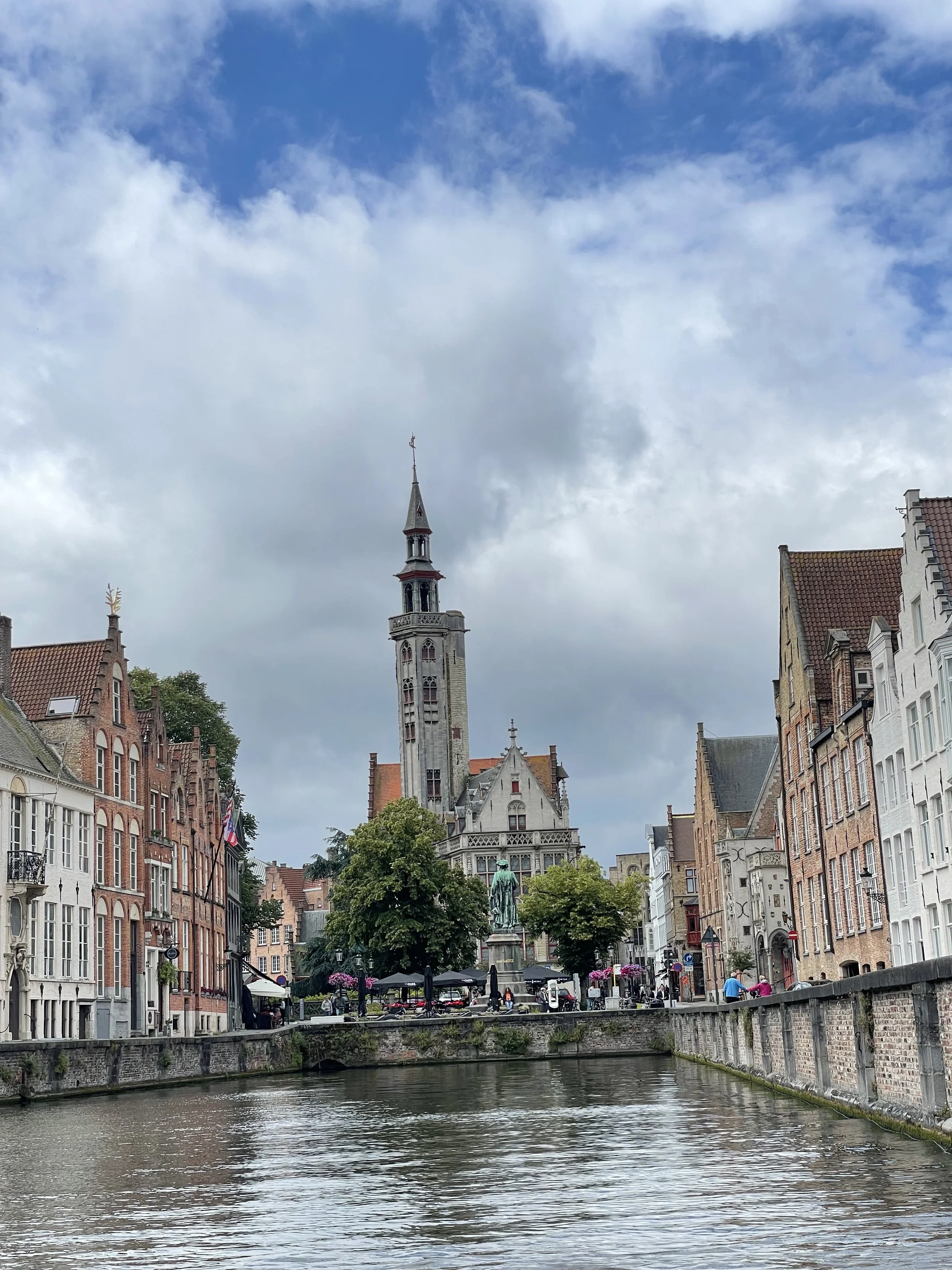 View of a city canal with historic brick buildings on both sides, a church tower in the background, and people walking along the street under a partly cloudy sky.