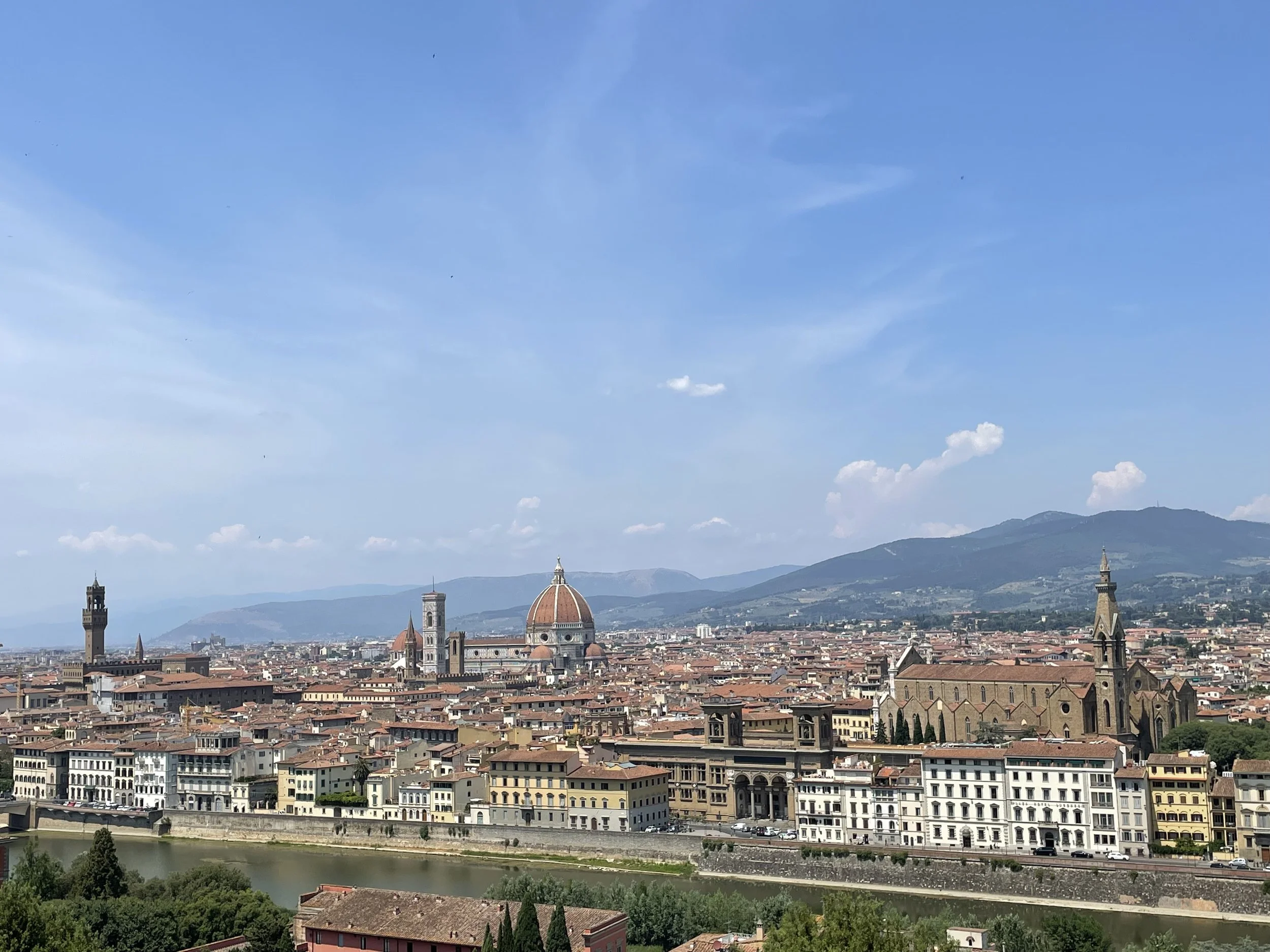 A panoramic view of Florence, Italy, with red-tiled rooftops, the Cathedral of Santa Maria del Fiore with its large dome, and the city skyline against a backdrop of mountains and a blue sky with some clouds.
