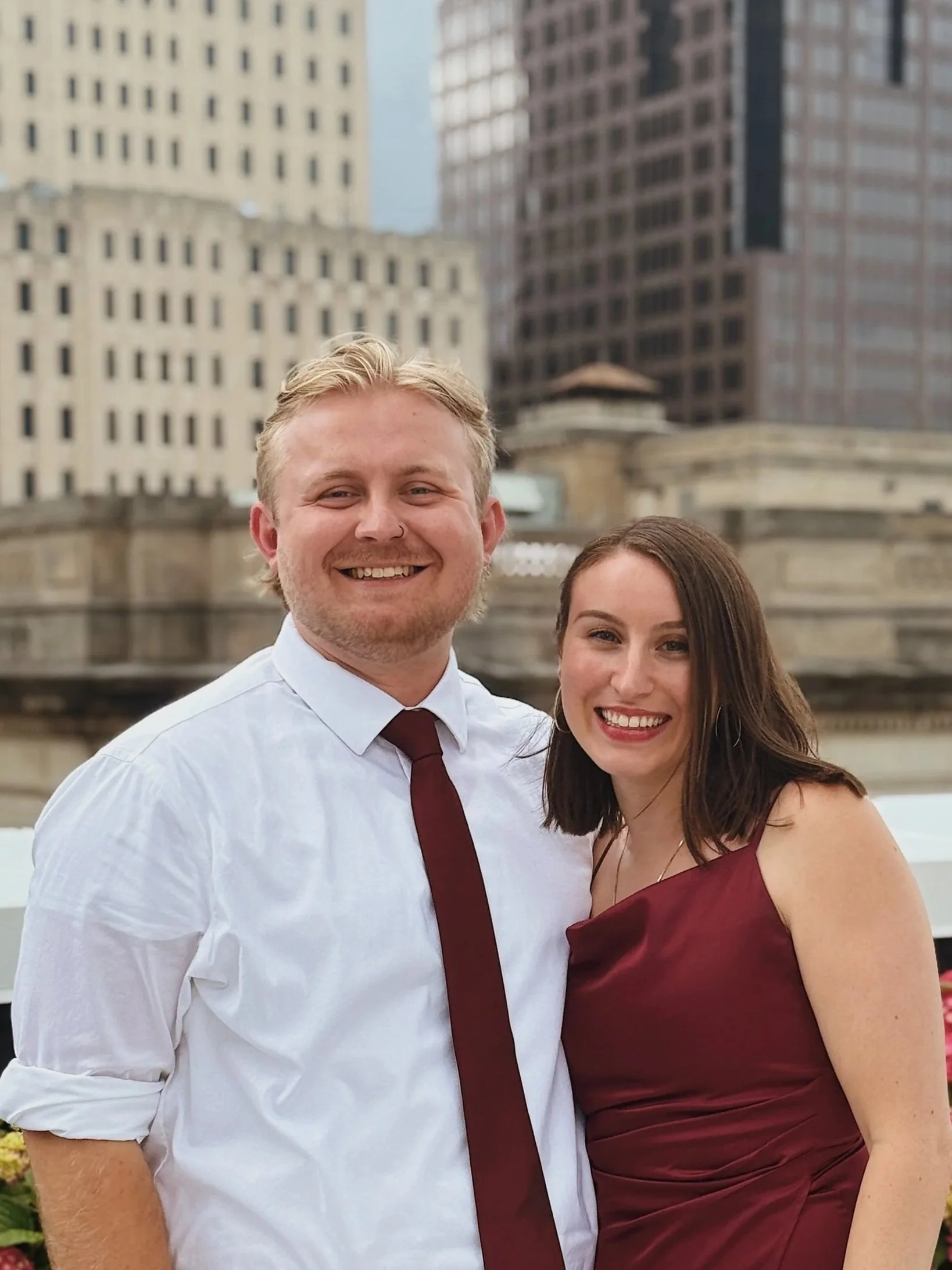A smiling young man with blond hair and a white shirt with rolled-up sleeves, and a smiling young woman with brown hair in a burgundy dress, standing outdoors in a cityscape with tall buildings in the background.