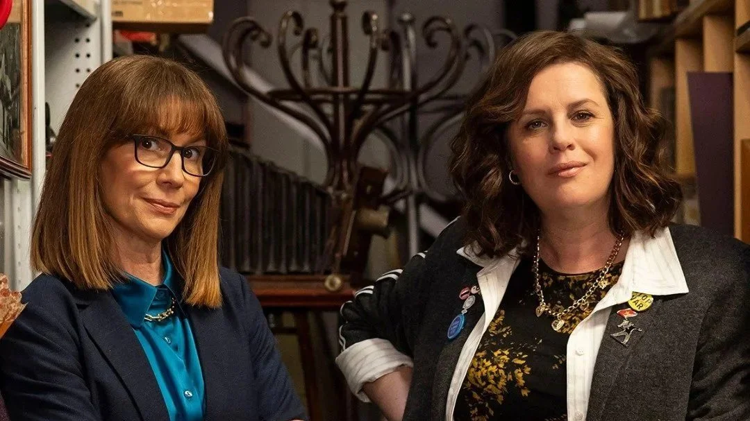 Two women standing in front of a collection of hats and objects, one with red hair and glasses, dressed in a blue shirt and blazer, the other with brown hair, wearing a dark blazer with pins and a chain necklace.