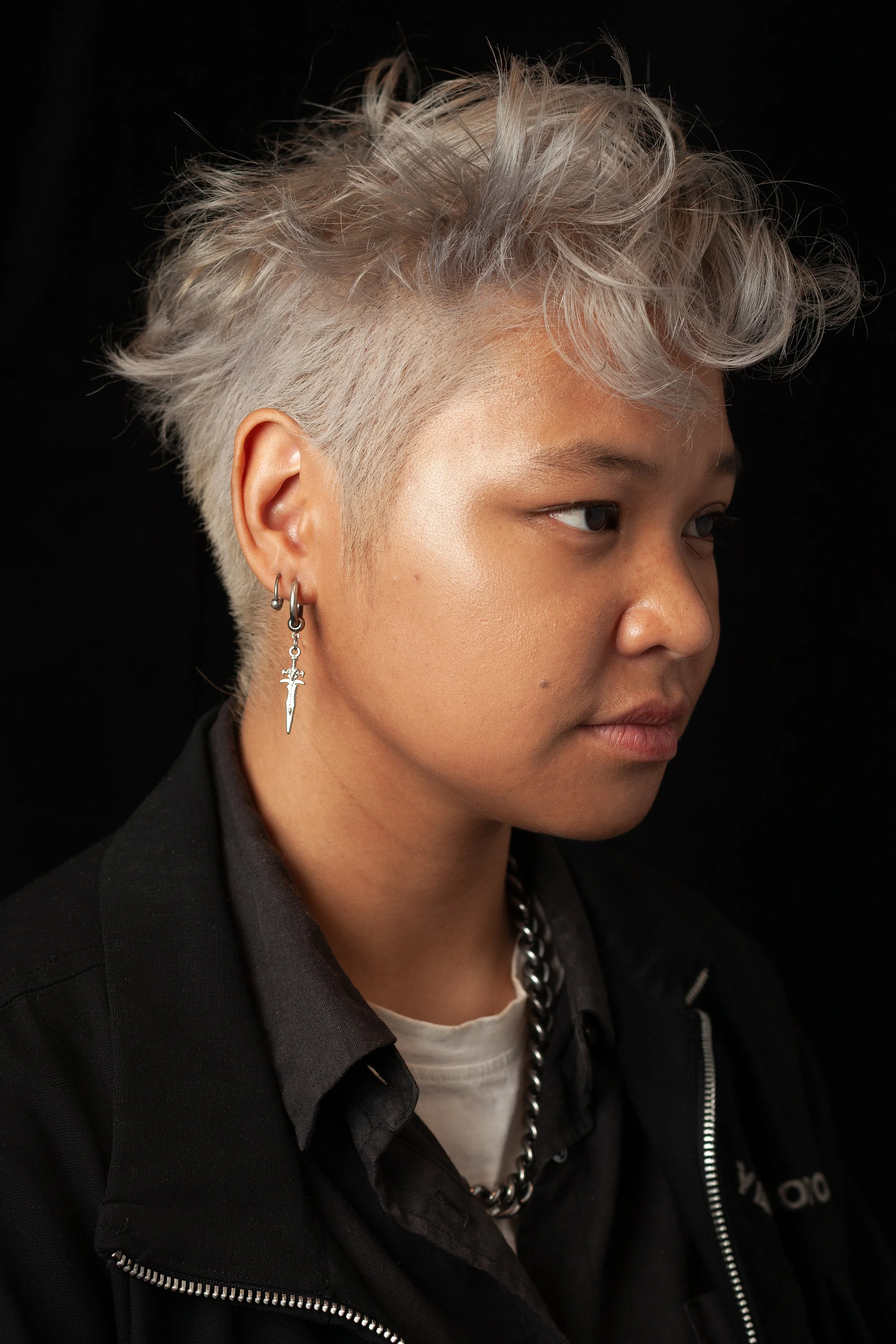 Portrait of a person with short, platinum blonde, messy hair, wearing earrings with swords, a chain necklace, and a black jacket against a black background.