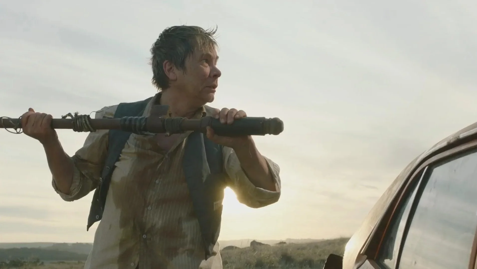 A man with disheveled hair and a dirty, beige shirt holding a large pipe staff on his shoulder, standing outside near a vehicle, with a landscape and cloudy sky in the background.