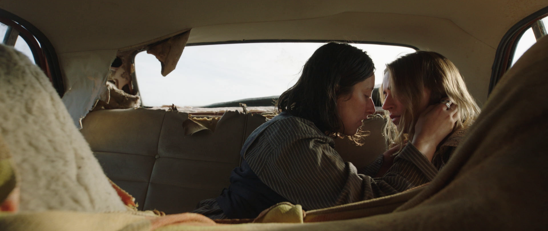 Two women sitting close together in the back of a worn, vintage car, gently touching foreheads with eyes closed, sharing an intimate moment.