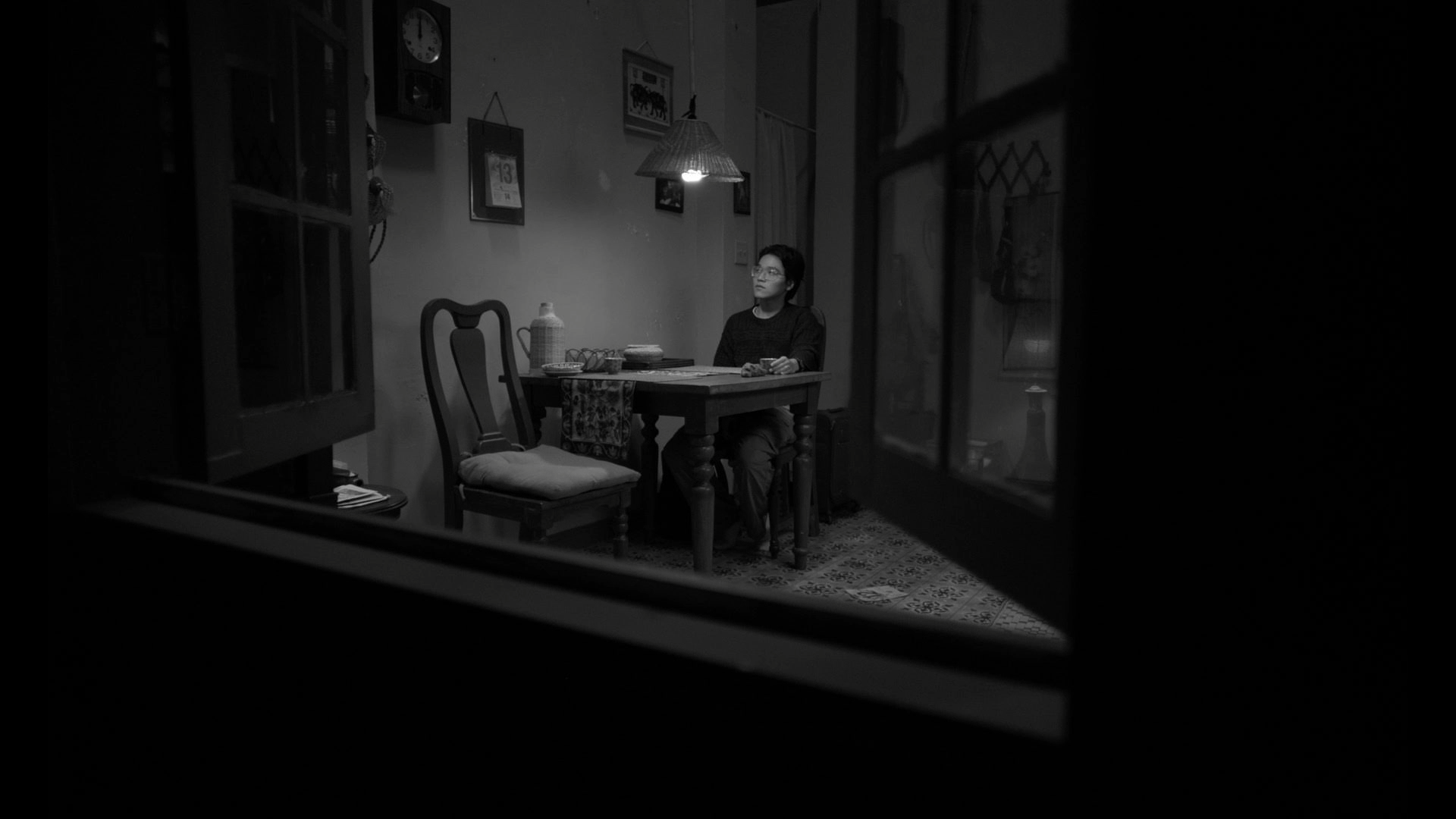 A woman sitting at a dining table in a dimly lit room, viewed through a window, with picture frames and hanging decorations on the wall behind her.