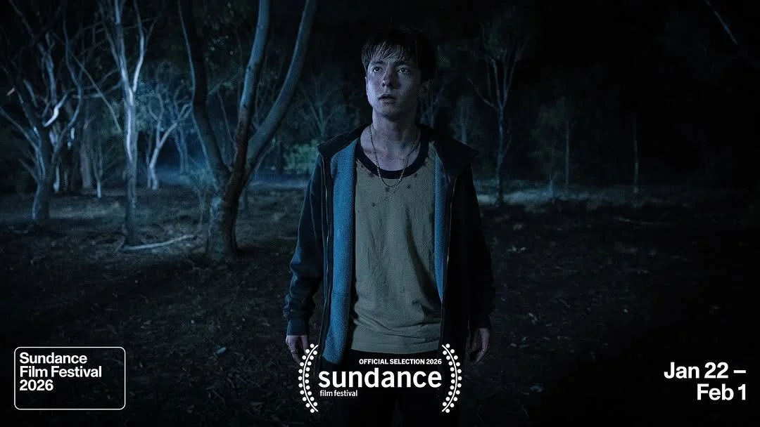 A young man stands alone in a dark, wooded area looking up with a concerned expression.