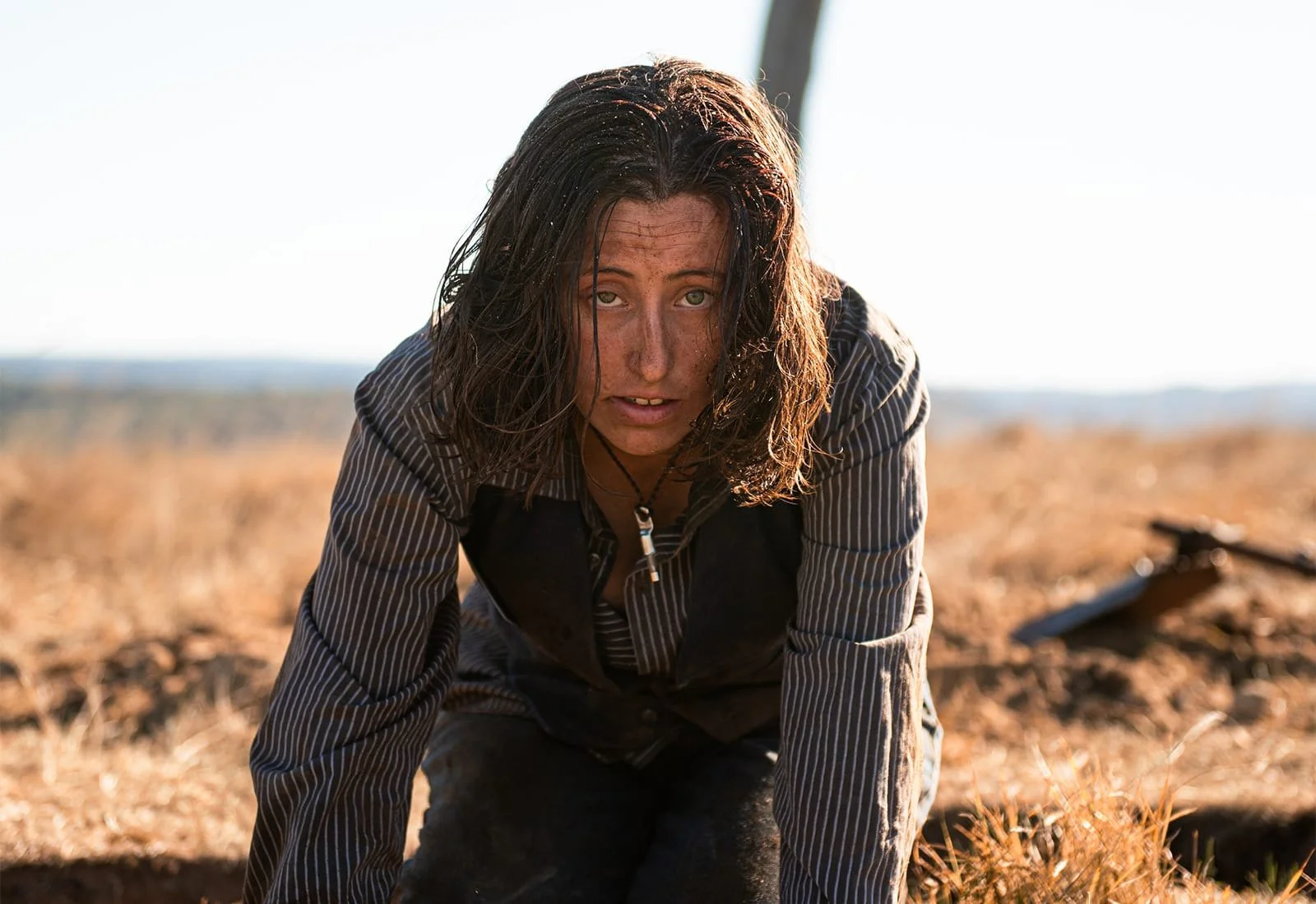 A woman with long messy hair, dirt on her face, wearing a striped shirt and a necklace, looks up while crawling on the ground in a dry outdoor landscape.