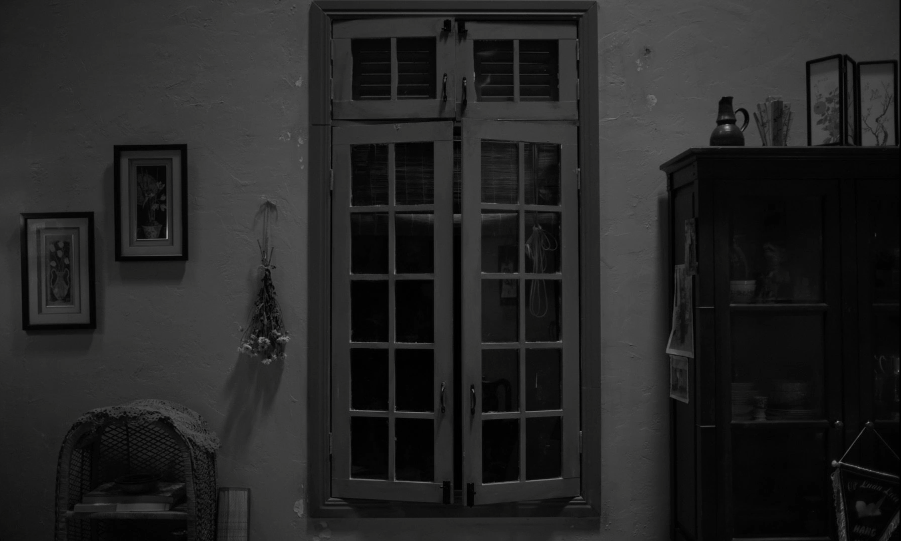 A black and white photo of an interior scene featuring a pair of closed window shutters in the center. To the left, there are three framed pictures hanging on the wall and a wicker chair with a lace cloth over the top, holding some items underneath. To the right, there's a tall dark cabinet with glass doors displaying dishes and decorative items. A small jute or string bundle with flowers is hanging on the wall beside the shutters.