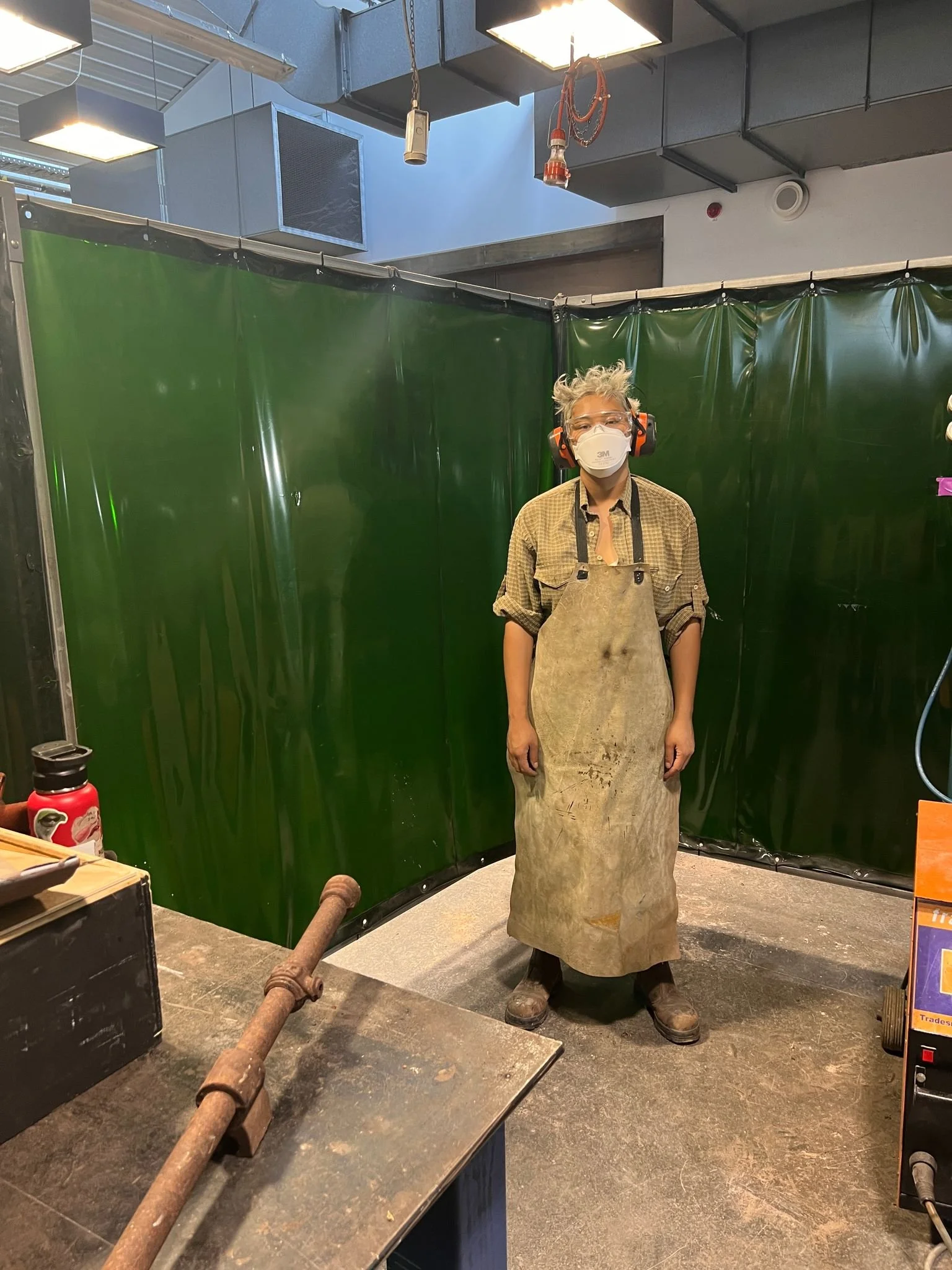 A person standing in a workshop or industrial setting, wearing safety gear including a face mask, ear protection, and a dirty apron. The background features green safety barriers, and there is equipment on the table nearby.