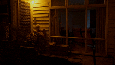 A child sitting on the ground outside a house at night, illuminated by warm yellow lighting.
